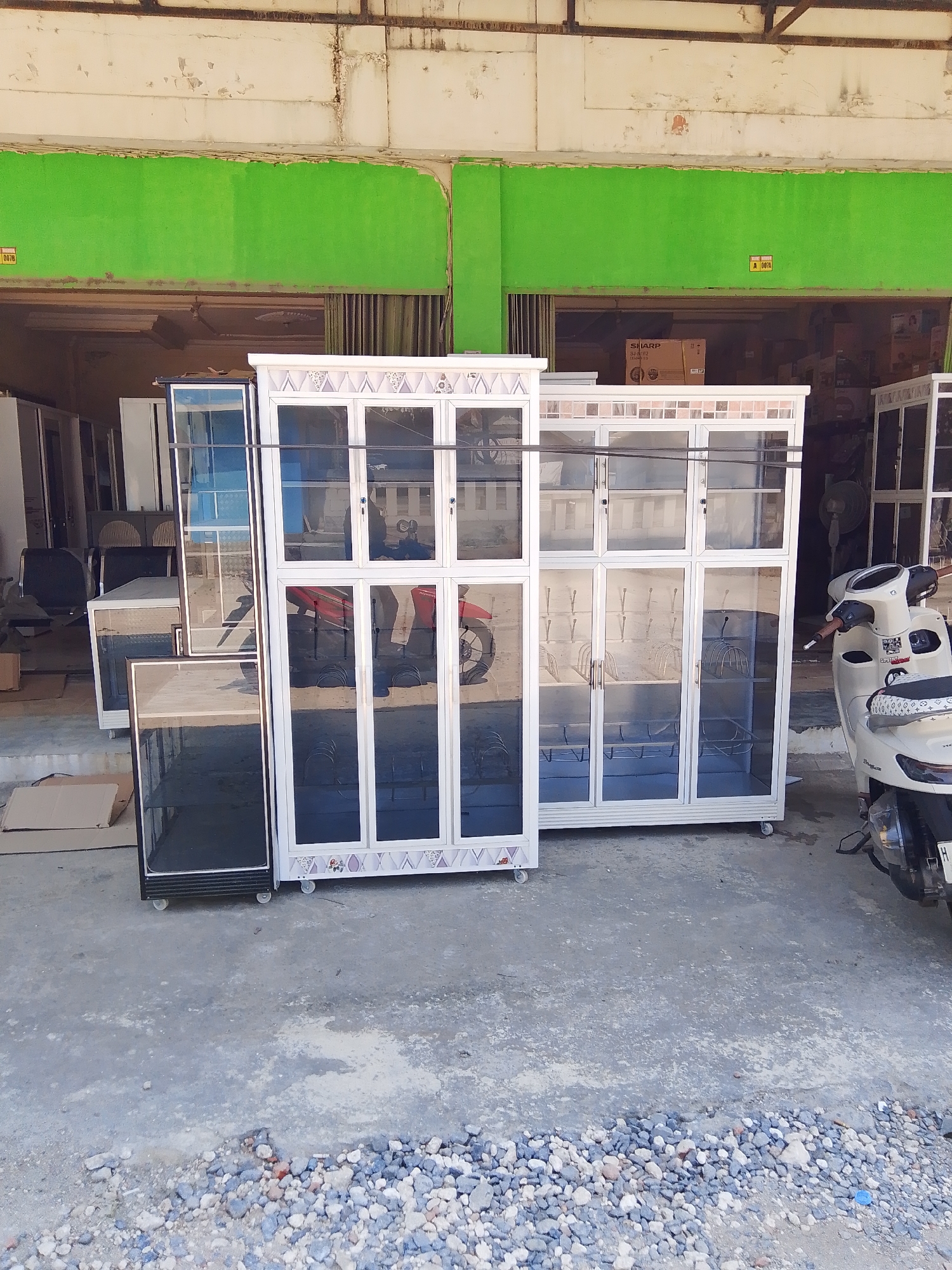 In Kedondong, Indonesia, an outdoor scene displays several tall, glass-fronted cabinets with aluminum frames, likely for display or storage, situated on a concrete ground. Two prominent cabinets are in the foreground, one to the left with a dark base and glass top, and a larger, white-framed cabinet to its right featuring intricate decorative tilework at the bottom and multiple glass doors. Inside the cabinets, metal racks and hooks are visible. A red motorcycle is parked behind the left cabinet, and a white scooter is parked to the right, with its rear visible. In the background, more cabinets and shelves are visible, along with some stacked cardboard boxes and what appears to be an open shopfront with a green painted wall. The overall environment suggests a commercial area or a shop selling furniture or household goods.