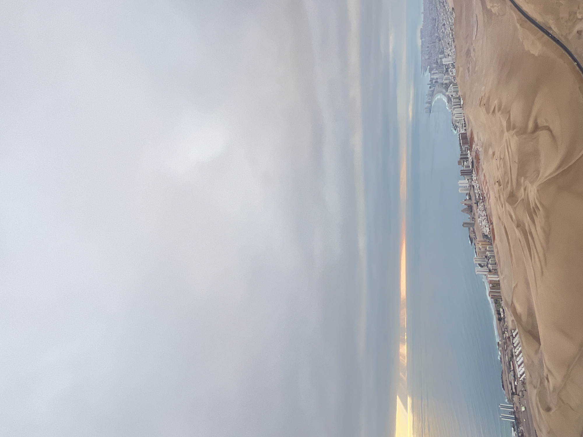 The image shows a view of a coastal city with a beach and buildings lining the coast, adjacent to a desert landscape under a cloudy sky. A road is visible near the desert region.