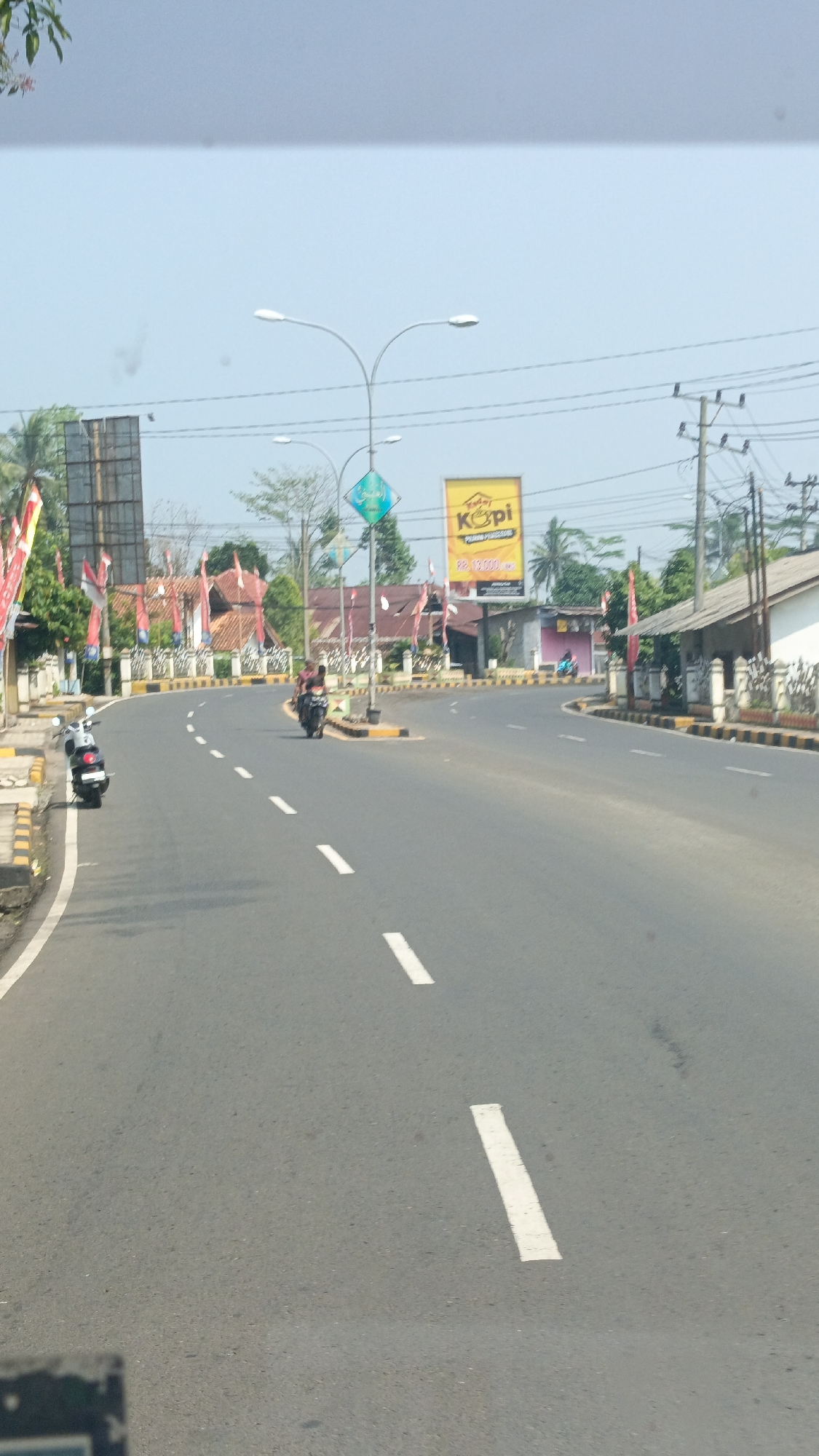 A highway in Kota Agung, Indonesia, features a motorcycle and scooter, both parked on the side of the road. The highway is lined with a sidewalk and a guardrail, separating it from the surrounding businesses and trees. A yellow billboard advertising "Kopi" stands out against the blue sky, with Indonesian flags displayed prominently on poles. A traffic light is visible in the distance, along with several power lines. The road is paved with asphalt and marked with white dashed lines, indicating a speed limit of 40 km/h. The scene depicts a typical day in a bustling Indonesian city, with its blend of modern infrastructure and traditional elements.