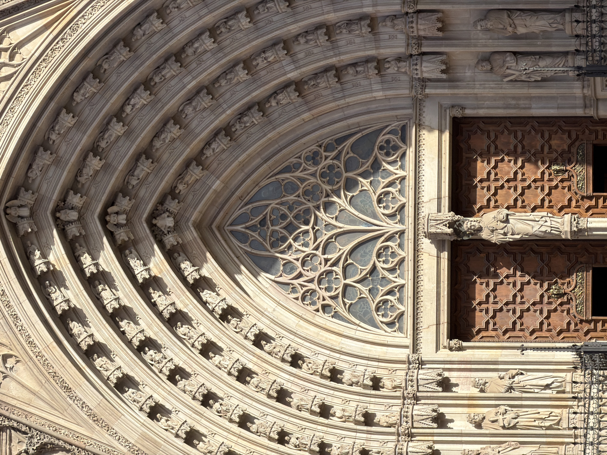 The image shows a detailed close-up of the main entrance of the Barcelona Cathedral, located in the Gothic Quarter of Barcelona, Spain. The entrance features a large, ornate Gothic archway with multiple layers of decorative carvings. Within the arch is a large, intricately patterned stained-glass window and a grand wooden double door. Flanking the doorway are several stone statues of saints and other religious figures, each standing in niches. The stone facade is weathered and shows signs of age, illuminated by natural sunlight casting shadows that accentuate the architectural details. The overall scene conveys a sense of historical grandeur and religious significance.