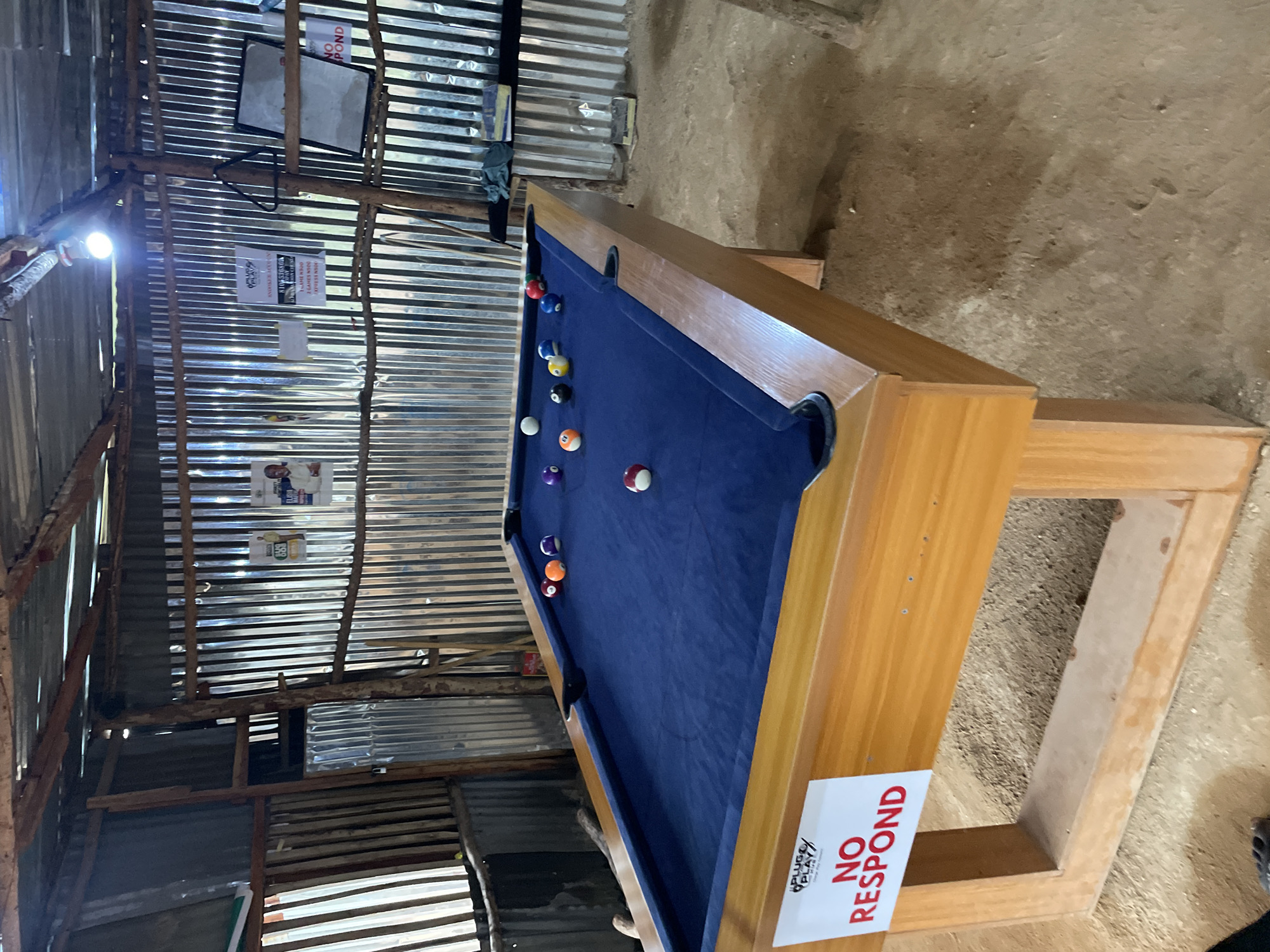 A pool table with a blue felt surface and a wooden frame is the central object in this media file. Several billiard balls are scattered across the table, including a white cue ball, a striped yellow ball, a solid purple ball, a solid orange ball, a solid red ball, and a solid blue ball. The table is situated in a rustic indoor setting with corrugated metal walls and a dirt floor. Wooden beams and supports are visible, suggesting a makeshift structure.
There are no people clearly visible in the image, nor is there an obvious activity or event taking place, though the presence of the pool table and balls implies a game of pool could be played. Notable details include a bright light fixture hanging from the ceiling, illuminating the scene. Several posters or notices are attached to the corrugated metal walls. One poster, partially visible in the top left, appears to be a white board with some writing. Another poster, located near the top center, is a white document with text. Further down, a poster features a picture of a person. On the side of the pool table, a sticker or sign with red and white text is prominently displayed, reading "NO RESPOND" along with a logo.
The overall impression of the scene is one of a basic, possibly outdoor or semi-outdoor recreational space in Durimi, Nigeria. The lighting suggests it could be daytime, but the interior setting makes it difficult to determine the exact time of day or weather conditions. The environment appears unrefined, with exposed materials and a dirt floor. The "NO RESPOND" sign might indicate a rule or instruction within the context of the location.