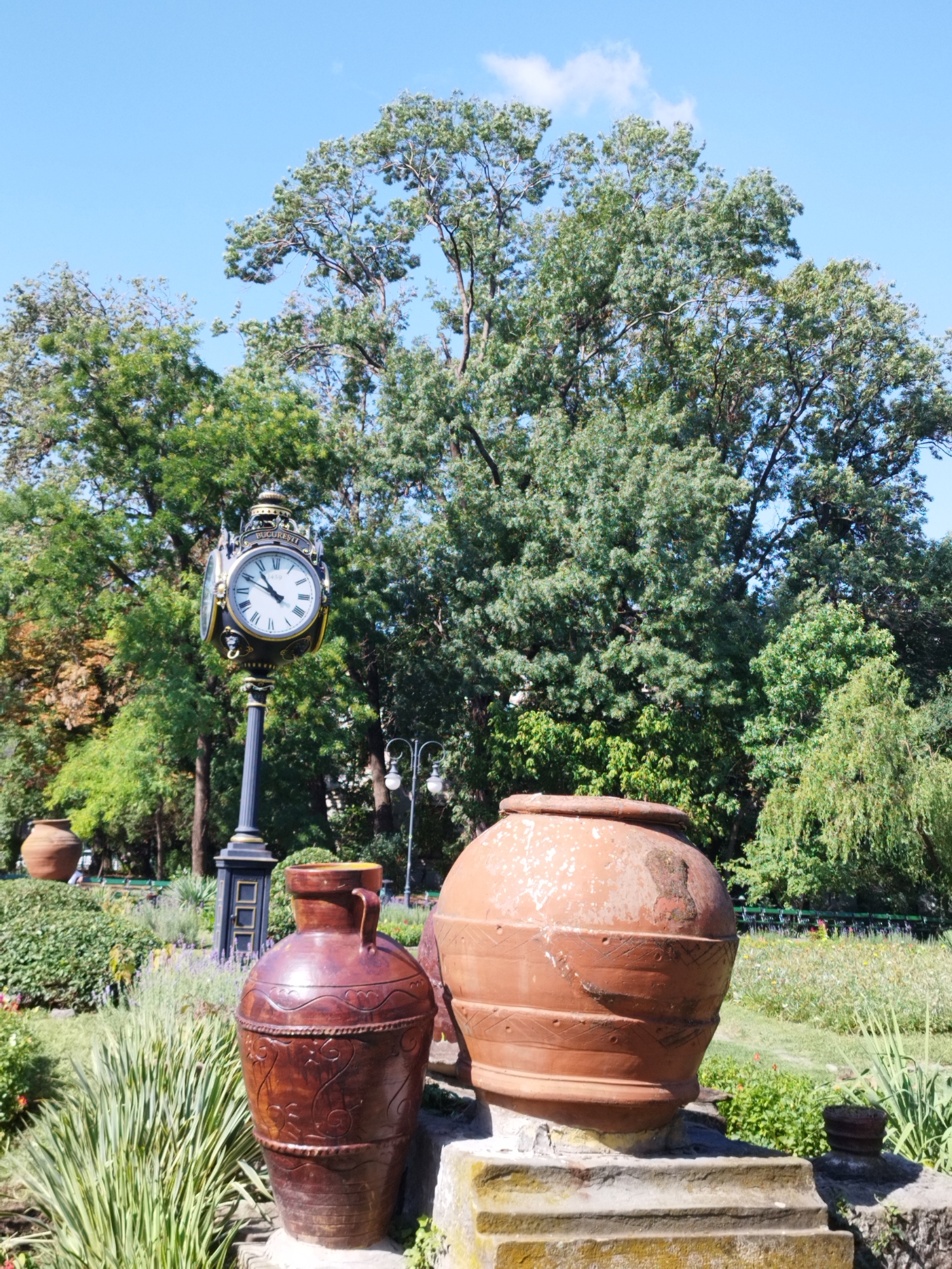 The image captures a sunny day in Bucharest, Romania, likely in a park or public garden. In the foreground, two large terracotta pots are prominently displayed. The pot on the right is a weathered, reddish-brown color with visible wear and tear, sitting on a stone pedestal. The pot on the left is a darker, glossy brown with intricate carved designs. Behind these pots, a classic street clock stands tall. The clock face is white with Roman numerals and ornate black hands, indicating the time is around 10:08. The clock is mounted on a dark, decorative pole with a base. In the background, lush green trees with dense foliage dominate the scene, with a clear blue sky above. The ground is covered with a variety of plants and flowers, suggesting a well-maintained garden. Some park benches are visible in the distance, hinting at a public space for relaxation and recreation. The overall impression is one of a tranquil and picturesque outdoor setting in the city.