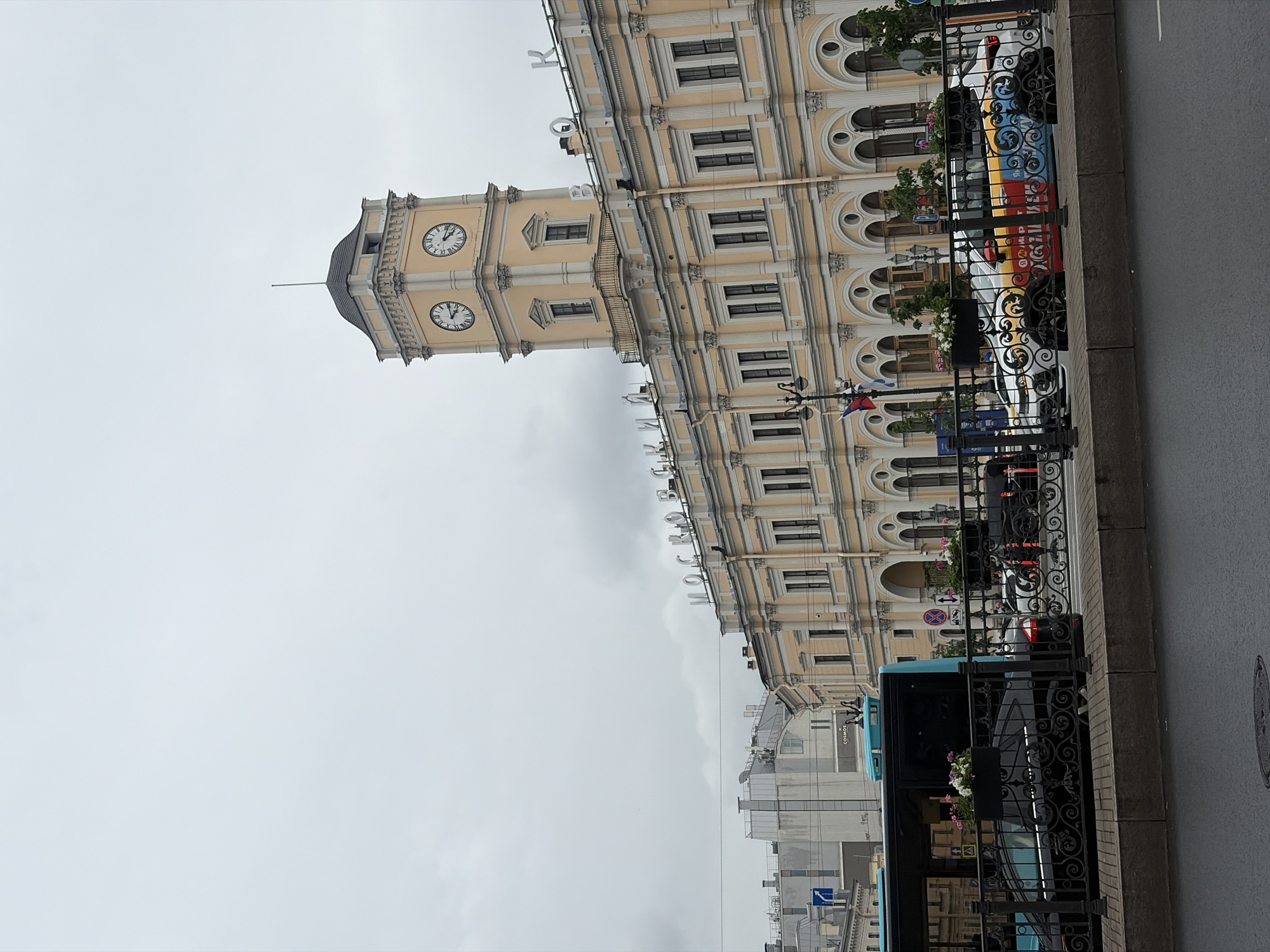 The image depicts Moskovsky railway station in Saint Petersburg, Russia, on an overcast day. The main subject is the station's prominent clock tower, which is pale yellow with dark trim and features two visible clock faces. The building itself is a large, pale yellow structure with multiple stories, adorned with arches and columns. Large, white lettering spelling "МОСКОВСКИИ" (Moskovsky) is visible across the building's facade, with individual letters extending upwards.
In the foreground, a black fence separates the sidewalk from the street. A blue bus is partially visible on the left, and several cars are parked behind the fence. One car is white with colorful, abstract graphics. Potted plants with pink and white flowers are placed on top of the fence. A street lamp with several flags is also visible. The street is wet, indicating recent rain. The overall environment appears to be an urban street scene.