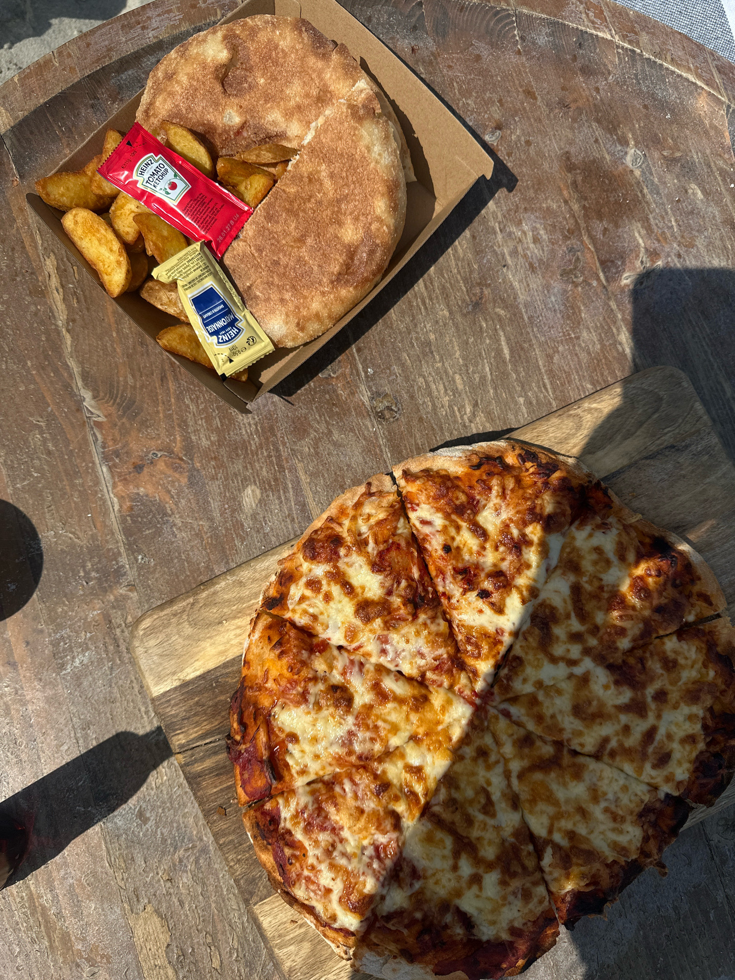 An overhead shot shows a pizza and potato wedges with condiments on a weathered wooden table, casting shadows from the sunlight. In the upper left of the image, a brown cardboard tray holds potato wedges and a circular flatbread. Two small packets, one red for Heinz tomato ketchup and one yellow for Heinz mayonnaise, are placed on top of the flatbread. To the right and lower down, a pizza, cut into six slices, rests on a wooden board. The pizza appears to be a Margherita with tomato sauce and melted cheese, with some charring on the crust. The overall scene suggests an outdoor dining setting.