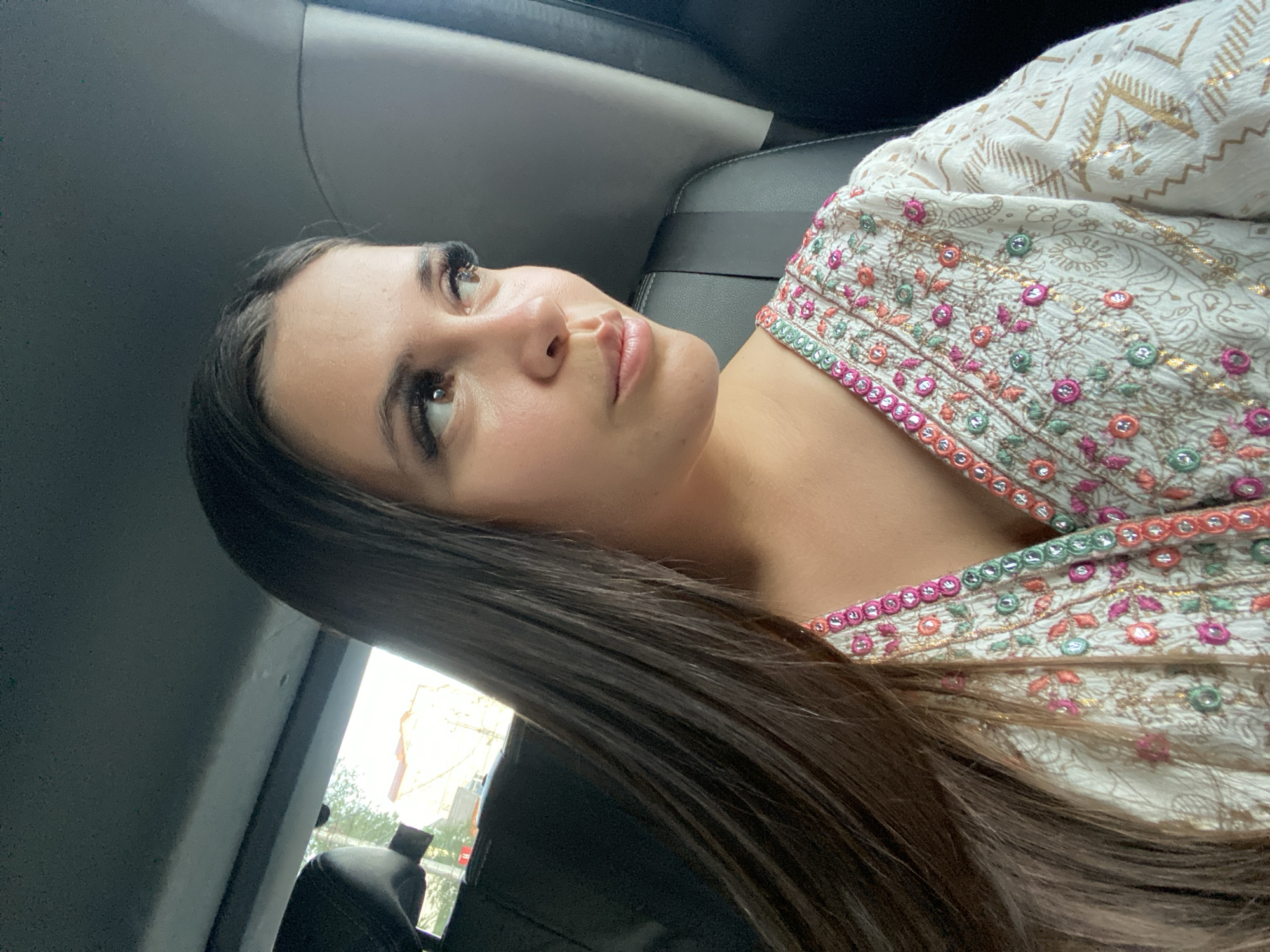 This is a portrait of a young woman in what appears to be the interior of a vehicle, possibly a car. She is looking upwards and to her right, with a neutral or contemplative expression. Her long, dark brown hair is visible, falling over her shoulder. She is wearing a white garment with intricate embroidery in various colors, including pink, green, and gold. The embroidery features floral and geometric patterns.
The setting is dimly lit, suggesting it might be daytime, but the lighting is not strong enough to determine the specific time of day. The background shows dark upholstery, consistent with the interior of a car. Outside the car window, there's a glimpse of a bright, possibly sunny, environment with some buildings and trees visible, hinting at an urban or suburban location. There is no visible text in the image. The overall mood is relaxed and personal, likely a candid shot taken within the vehicle.