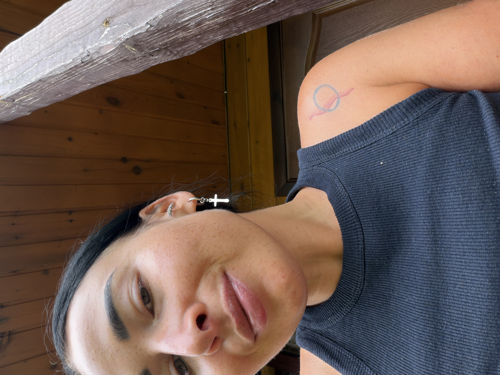 A close-up, low-angle, indoor selfie shows a woman with dark hair and dark eyebrows, wearing a black ribbed tank top. She has freckles on her face and a small, silver cross earring in her left ear. A tattoo is visible on her right shoulder, appearing as a pink stroke inside a blue circle. The background consists of wooden planks, suggesting an indoor setting possibly in Sochi, Russia, as hinted by the wooden structure.