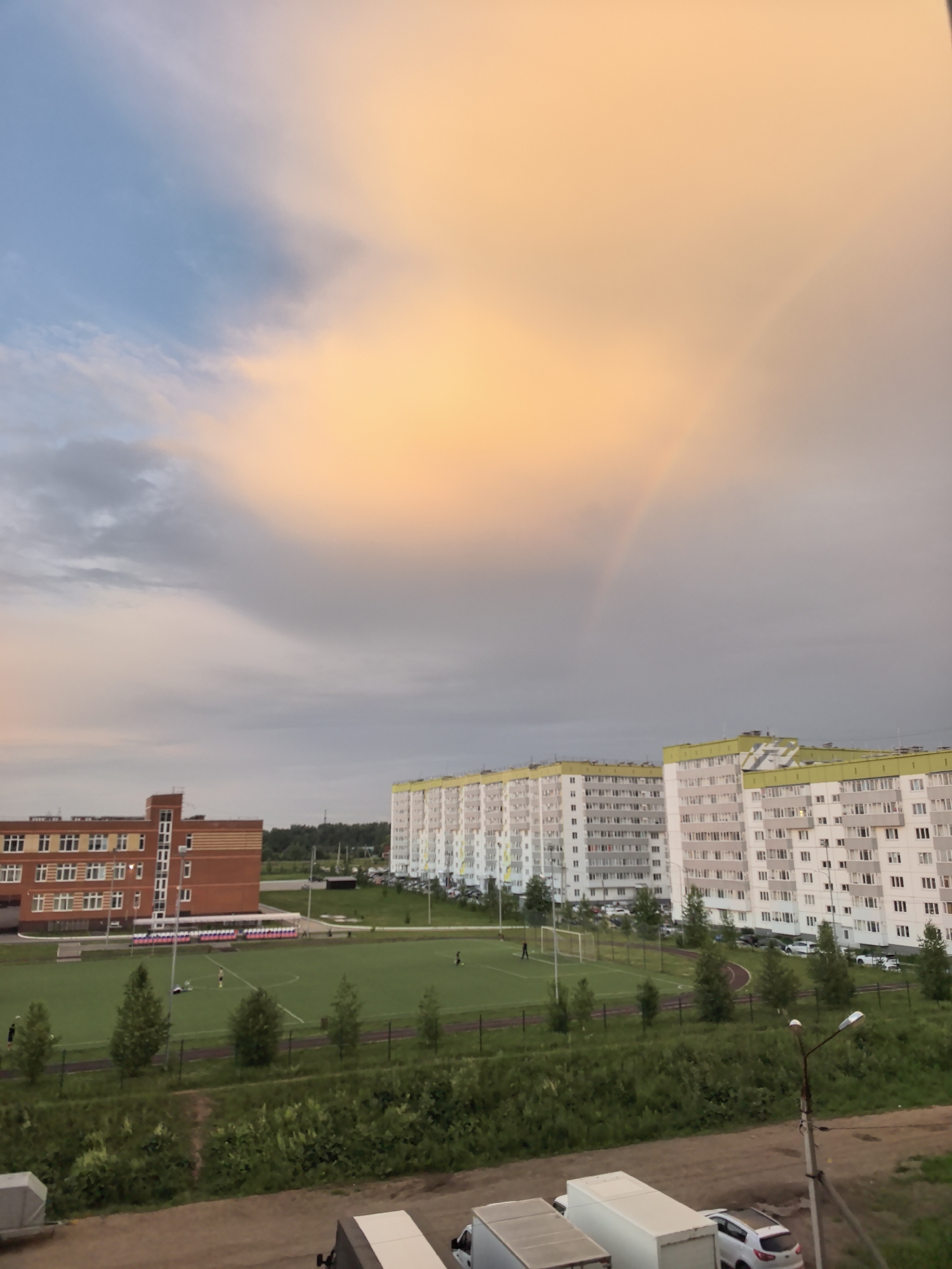 The image depicts an outdoor scene in the city of Фролы, Russia, featuring a football field, residential buildings, and a cloudy sky with a rainbow. The football field is green with white markings and a goalpost visible on the right side. Several people are present on the field, some appearing to be playing football, while others are walking or standing. To the left of the field is a brick building with multiple windows, resembling a school or administrative building. Two tall, white residential buildings with yellow trim are situated behind the football field. The sky is a mix of blue and orange clouds, with a faint rainbow arching across the upper right portion of the image. Trees and green vegetation are scattered around the field and buildings. In the foreground, a dirt road is visible, with parked trucks and cars. The overall impression is a suburban or semi-urban environment during a cloudy, possibly post-rainy, day.
