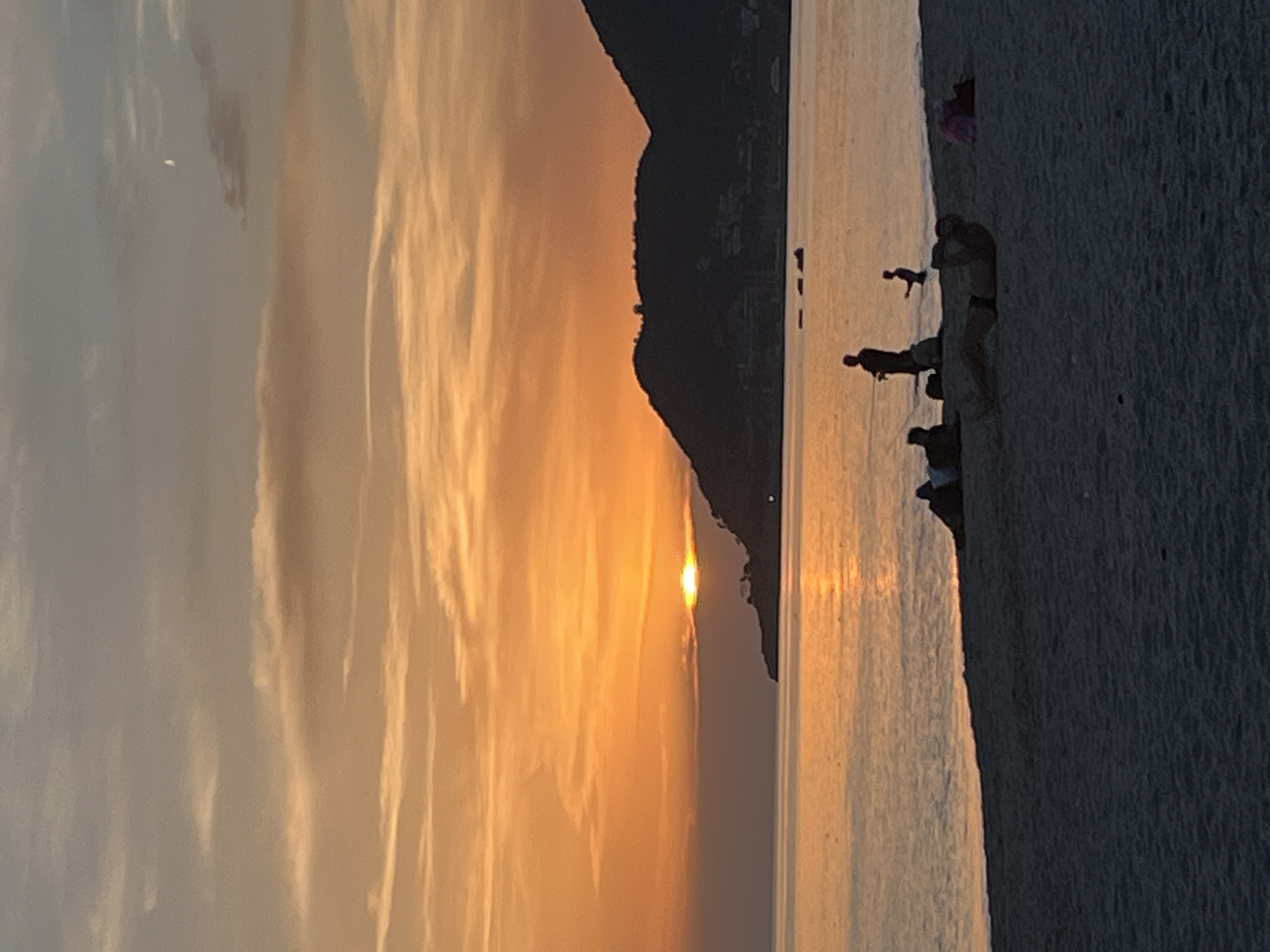 This photograph captures a sunset scene in Alanya, Turkey. The sky is a blend of orange and yellow hues with streaky clouds. A dark, silhouetted mountain or landmass is positioned near the center. The sea reflects the sunset's colors. Several silhouetted figures stand on the beach, near the water's edge. The sand is visible.