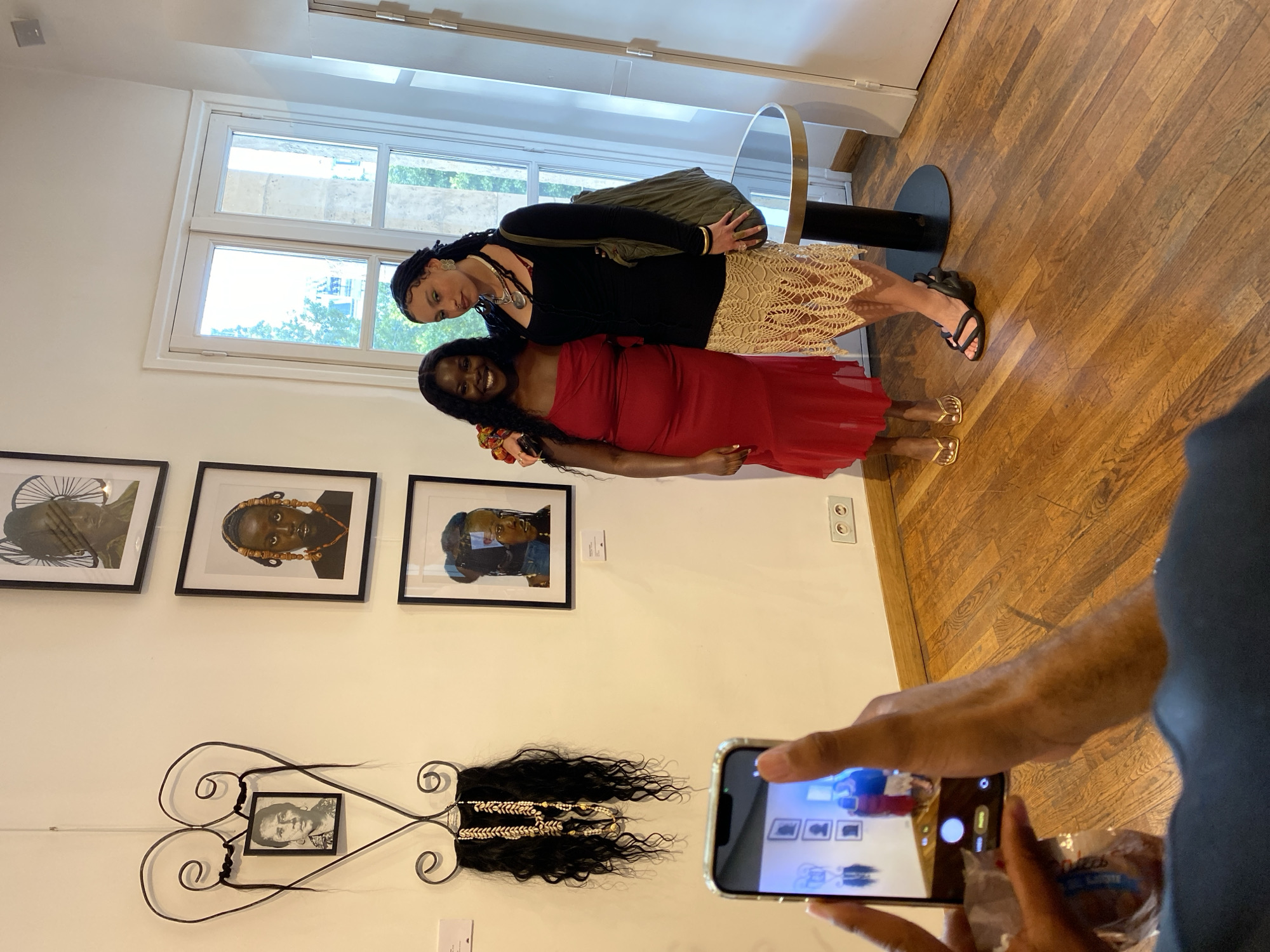 Two women are posing for a photo in front of an art display in what appears to be an art gallery. A person holding a smartphone is taking their picture. The display includes three framed photographs and an art piece featuring a black and white photo, metal swirls, and a section of black hair decorated with beads. The location appears to be in Paris, France. The floor is hardwood, and a window is visible in the background.