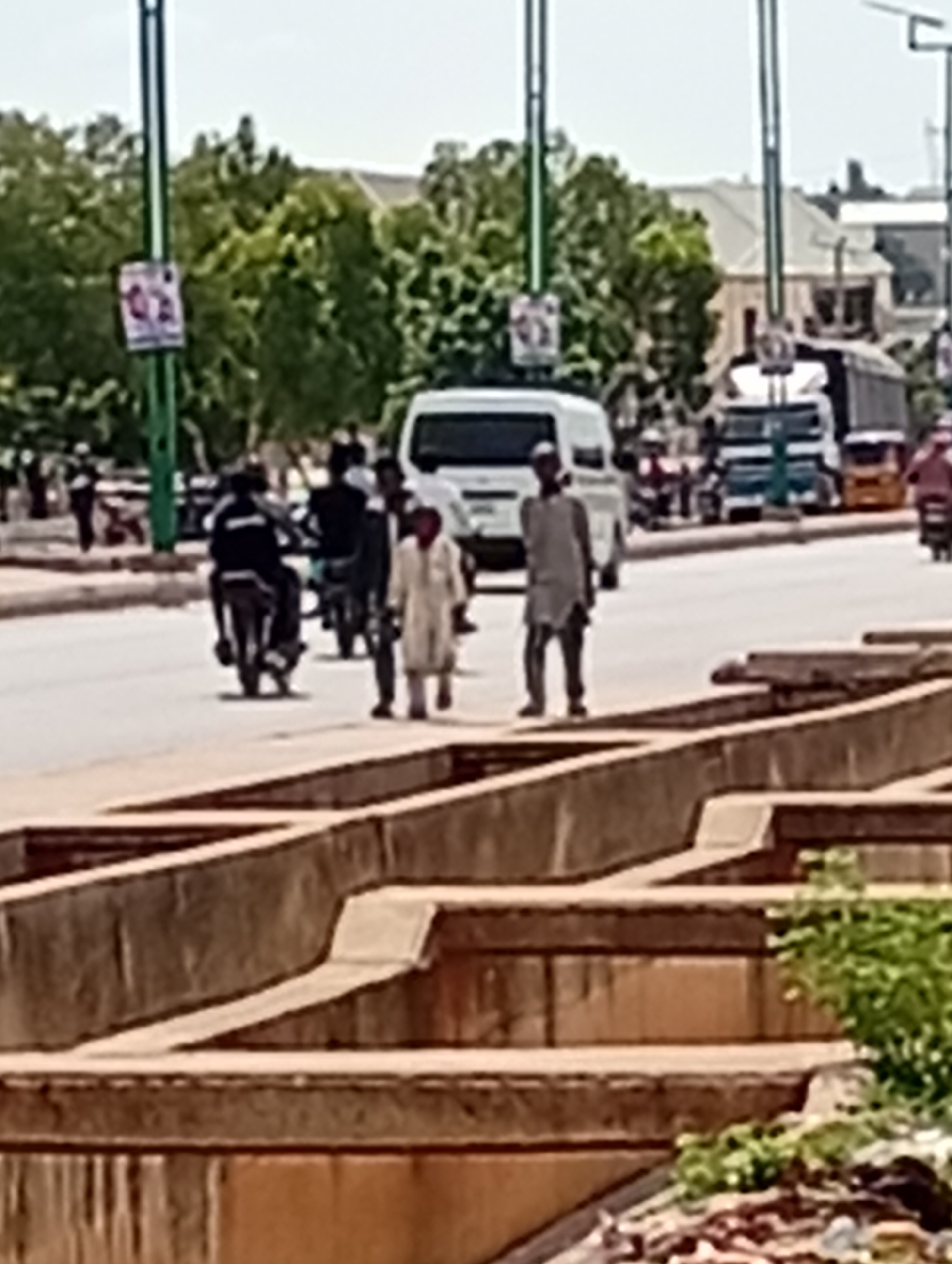 The image captures a street scene in Tumfure, Nigeria, under a bright sky. In the foreground, a concrete structure with multiple levels and a partially visible drainage system is prominent. On the street, two men are walking, one wearing a light-colored, traditional robe and the other in darker clothing. A white van is parked behind them, and a motorcycle is in motion to the left, with its rider obscured. Further down the road, several other vehicles, including trucks and what appears to be a tricycle taxi, are visible, along with more people and trees lining the street. A lamppost with a sign is also visible on the left side of the image.