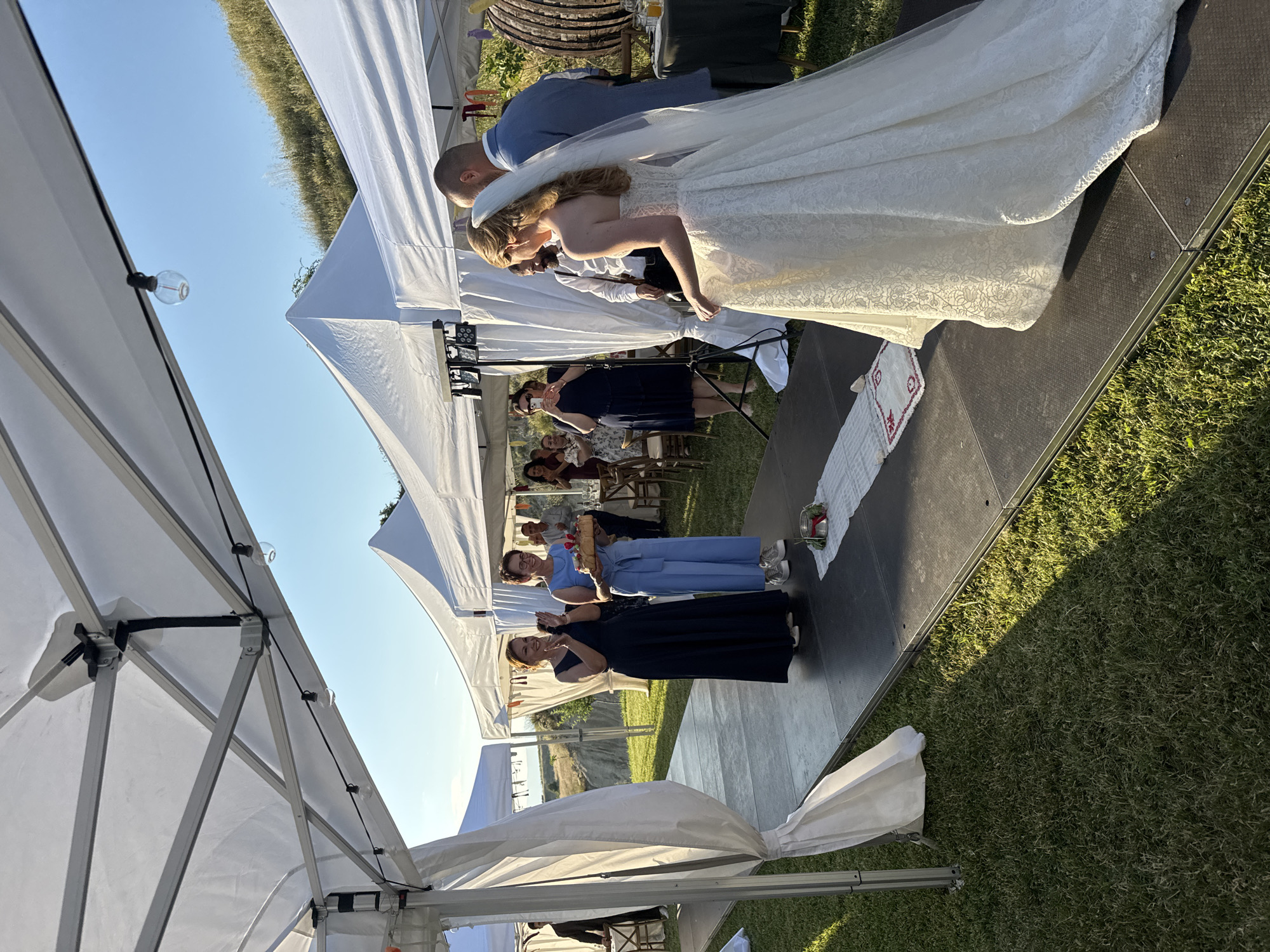 The image depicts an outdoor wedding event in Harsovo, Bulgaria, set on a grassy area under multiple white canopy tents during daylight hours with clear blue skies. In the foreground, a bride in a white strapless lace gown and a long veil walks on a raised black modular platform, with her back mostly to the camera, accompanied by a man in a light blue suit jacket.
To the left on the same platform, two women stand, facing the approaching couple. One woman, wearing a dark blue dress, is clapping and smiling. Beside her, a second woman, wearing a light blue jumpsuit and glasses, holds a large, light-colored object, possibly a ceremonial bread or cake, decorated with red elements. On the platform in front of these women, a white fabric runner with red embroidered patterns and a small silver bucket containing greenery and red berries are visible.
Several guests are visible in the background under and around the tents. One woman in a dark dress is seen taking a photograph with a smartphone. Other guests are seated at tables with wooden chairs or standing, observing the scene. A light stand with multiple rectangular fixtures is positioned behind the woman taking a photo.
The tents feature visible metal frame structures, from which string lights with clear bulbs are suspended. White fabric drapery is tied back on the left side of the foreground tent entrance. The ground around the platform is covered with green grass, exhibiting shadows cast from the tents and individuals, indicating direct sunlight from the right side of the frame. In the far background, rolling hills covered in dry grass and vegetation are visible under the clear sky, with a more distant eroded landscape present on the left. On the far right, a large, barrel-like wooden structure and a dark-colored table with beverages and glasses are present under a tent.