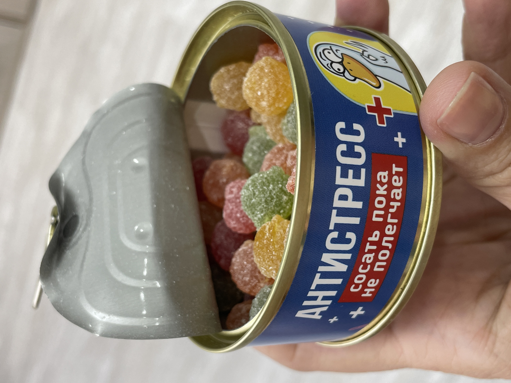 A close-up shot features an open tin can being held in someone's hand. The can, labeled "Антистресс" (Antistress) in Russian, with the tagline "Сосать пока не полегчает" (Suck until it gets easier), contains multicolored, sugar-dusted gummy candies. The can's label also displays a cartoon-style goose graphic in the upper right corner. The background is a light-colored surface. Based on information available online, "Antistress" candies in such tins are sold in Izhevsk, Russia.