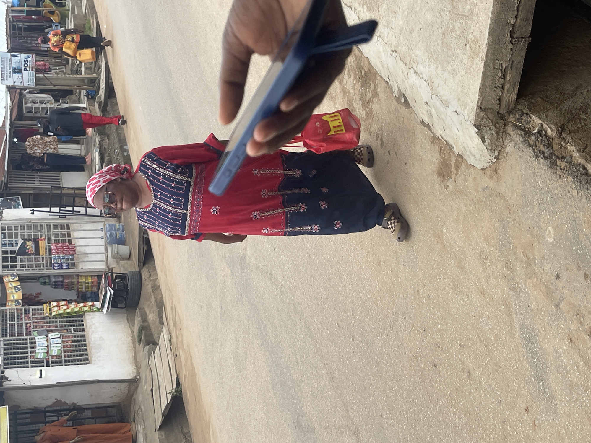 This image captures a vibrant street scene, likely in a commercial area of Abuja, Nigeria, during daylight hours. The perspective is from slightly above, looking down a paved street.
In the mid-ground, a woman is the central subject. She wears a long, flowing garment with a red top section featuring intricate blue and white patterns, and a dark blue lower section with similar patterns. A red and white patterned headscarf covers her head, and she wears eyeglasses. A red shopping bag is slung over her shoulder. On her feet are distinctive checkered slip-on shoes, likely Crocs. Her expression is neutral as she appears to be walking.
In the immediate foreground, a dark-skinned hand holds a blue smartphone, angled downwards as if taking a photo or recording. This hand partially obscures the woman and the ground directly in front of her.
The background to the left reveals a bustling street with several people and small businesses. A person in a black top and red trousers walks away from the camera. Further down the street, another individual wearing an orange vest is seen carrying two yellow jerrycans. The shops lining the street feature metal grilles and display various goods, including packaged food items and possibly paint cans. Visible text on signs includes "POS" (Point of Sale) and "LUKELAND COLLECTIONS."
To the right, a weathered concrete wall or barrier runs parallel to the street, showing signs of wear and tear with exposed aggregate and rough edges at its base. The ground throughout the scene is a light-colored, somewhat textured paved surface, consistent with a road or walkway. The overall atmosphere suggests a typical day in a busy urban environment, with people engaged in daily activities.