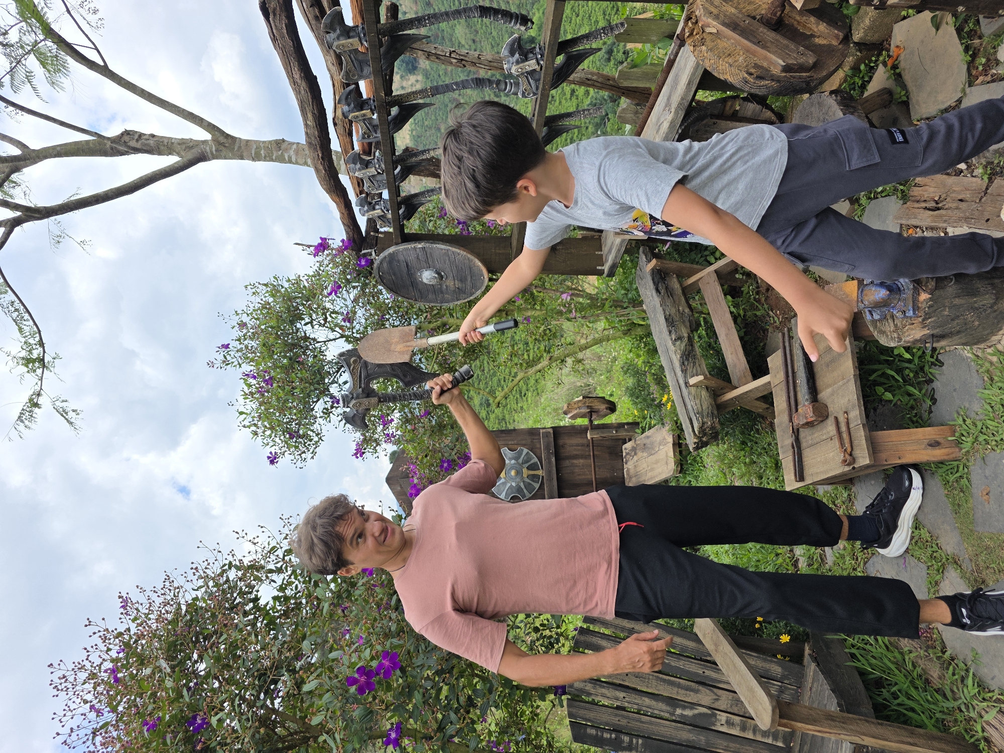 The image, taken in Thôn 1, Vietnam, features two individuals amidst a rustic, possibly themed, outdoor setting. A young boy, wearing a gray t-shirt and dark pants, extends his hand toward an axe held by a man in a light pink t-shirt and black pants. The man appears to be in mid-motion, possibly posing with the axe raised above his head. Behind them, a display of various axes and a shield is arranged on a wooden structure. The background includes greenery, likely natural foliage, and partially visible wooden benches. The scene appears to be staged or part of a display within Thôn 1.