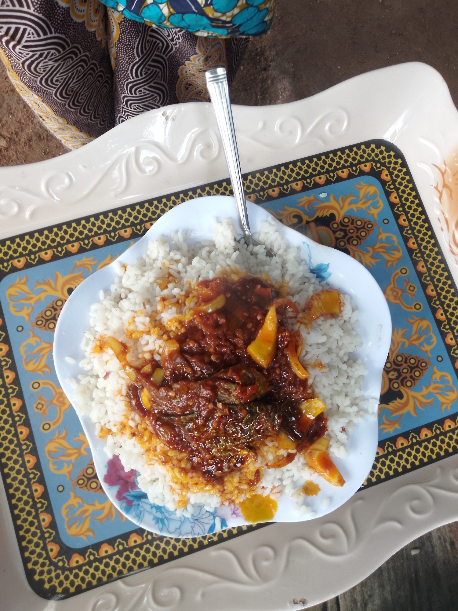A plate of white rice topped with a reddish-brown stew containing fish and yellow-orange pieces, likely peppers or sweet potatoes, is positioned on a decorative blue and gold tray. A silver fork rests on the plate. The tray is situated on a white, embossed platter, which is placed on a surface of dark brown material, possibly wood or dirt. In the upper left of the image, the patterned fabric of clothing, in shades of brown, black, and blue, is visible, suggesting a person is present but not fully in frame. The scene appears to be an outdoor setting, potentially in Serti, Nigeria, as implied by the context.