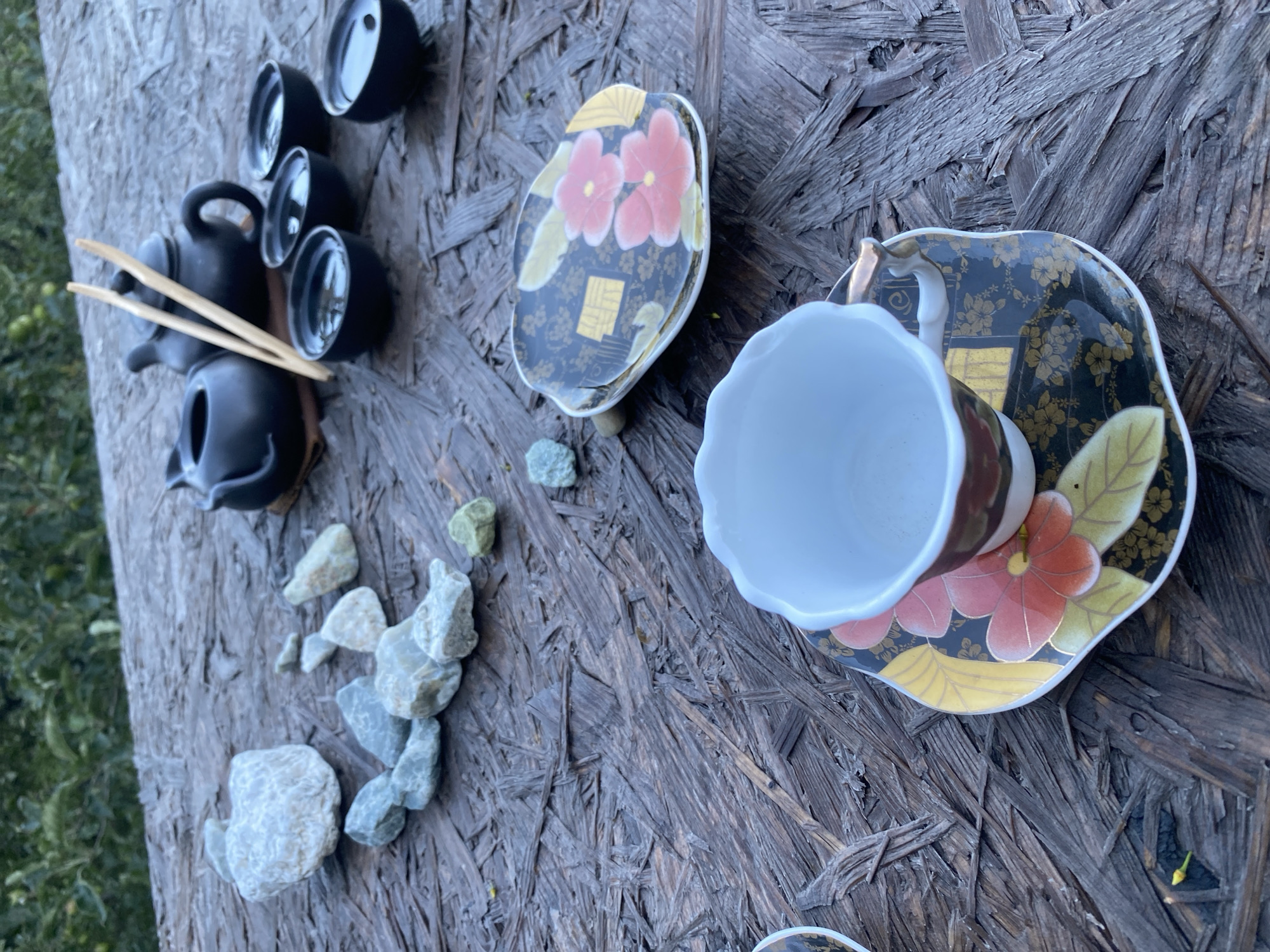 The image depicts an outdoor tea ceremony setup on a weathered wooden surface, possibly a table. The background suggests a natural setting with greenery visible, hinting at a park or garden environment. In the foreground, an empty teacup sits on a saucer adorned with floral patterns in red, yellow, and gold against a black background. Beside it, another decorative saucer is partially visible. Further back, a small black teapot and several small black tea bowls filled with liquid are arranged. A pair of wooden chopsticks rests atop the teapot. Scattered around the setup are several light-colored stones, some appearing smooth and rounded, while others are more irregular in shape. The overall scene conveys a sense of calm and tradition. There is no indication of any specific location within the city of Almaty, Kazakhstan.