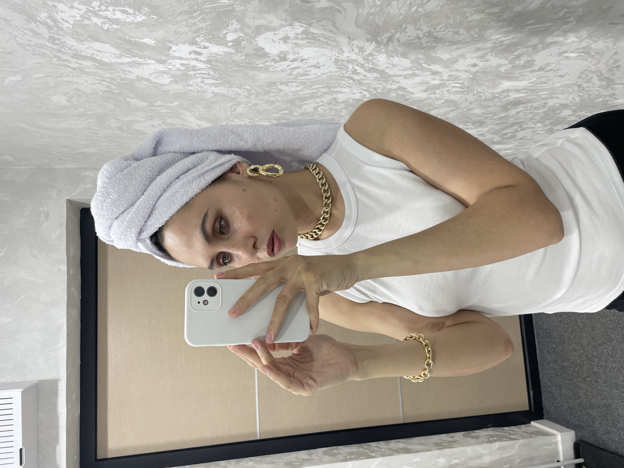 A woman in a white tank top and a towel wrapped around her head is taking a selfie in a mirror. She is wearing a gold chain necklace, earrings, and bracelet. The mirror reflects a bathroom setting with a gray and white marble counter and a black-framed mirror. A cabinet is visible on the left wall, and the ceiling is painted with a swirled pattern. The woman holds a white smartphone to take the photo. There is no information about the location in the city of Izhevsk, Russia.