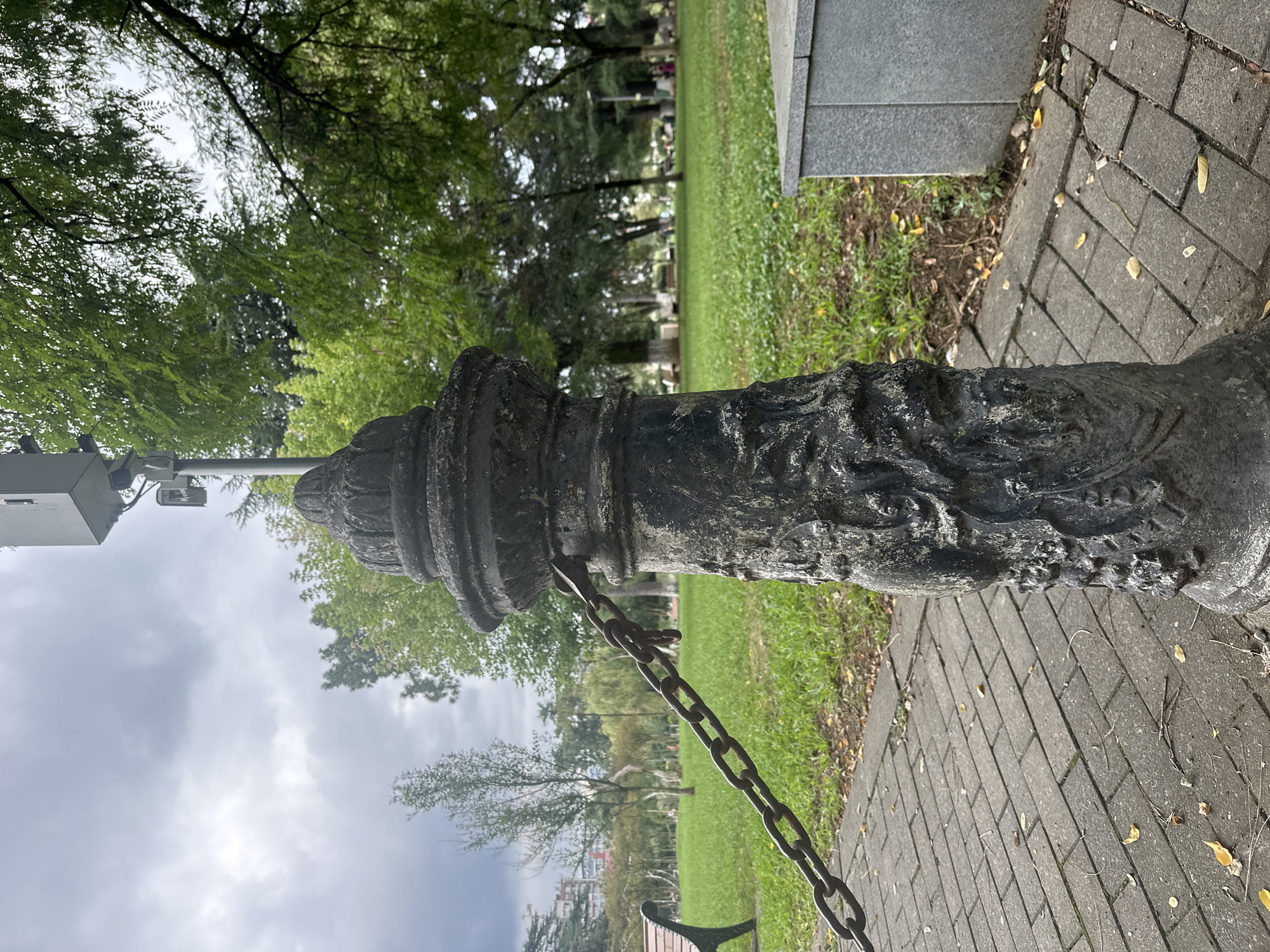 The image shows a decorative metal bollard with a chain attached to it, located on a paved pathway in a park. The bollard is weathered and appears to be made of cast iron, featuring intricate carvings. A heavy metal chain connects the bollard to another unseen object, suggesting it might be part of a barrier or enclosure.
In the background, a lush green lawn stretches out, dotted with trees and park benches. The sky is overcast, with dramatic clouds suggesting a changing weather. In the distance, buildings can be seen, indicating an urban setting.
This scene is located in Batumi, Georgia, a city known for its beautiful parks and coastal promenade. The specific location appears to be part of a public park, possibly along the Boulevard or a similar green space within the city. The presence of the bollard and chain could indicate a demarcation of an area, a decorative element, or a way to control access. There are no people visible in the image. The overall impression is one of a tranquil, if slightly moody, park environment on a cloudy day.