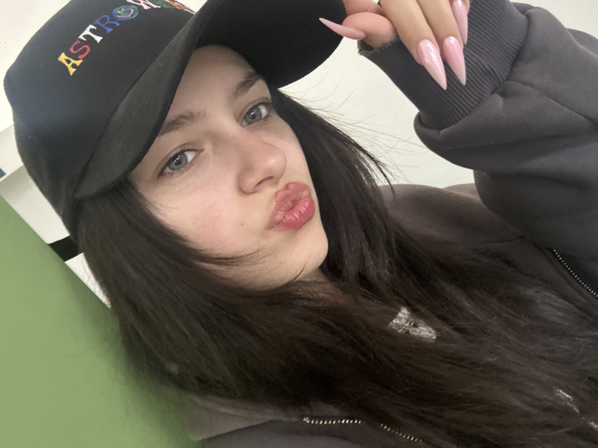 A person is posing for a close-up selfie. The person is wearing a black baseball cap with the word "ASTROWORLD" embroidered in colorful letters, and a gray jacket. The person has long, dark hair and light eyes, and is puckering their lips. They are holding their hand up near their hat, showing long, pointed, pink nails. The background is indistinct, suggesting an indoor environment with pale green walls. There is no information to suggest a location in Yerevan, Armenia.