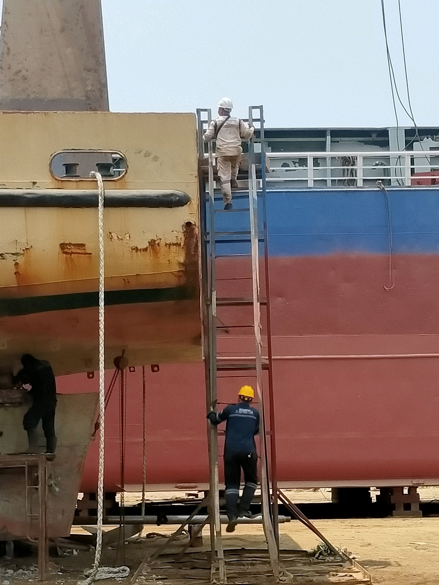 In Kamal, Indonesia, a dry dock facility is depicted with several workers engaged in maintenance on a ship. In the foreground, a worker in a dark blue uniform and yellow hard hat is climbing a ladder attached to the side of a large vessel. The vessel has a distinct two-tone paint scheme, with the upper section painted blue and the lower section a deep red. To the left, another worker, dressed in black, is positioned on a platform near the ship's hull, appearing to be working on the lower part of the structure. Further up the ladder, a third worker, wearing a light-colored uniform and white hard hat, is ascending, approaching the upper deck of the ship. The ship's hull exhibits signs of wear and rust, particularly on the older, yellow-painted section on the left. A thick rope is hanging vertically along the side of the yellow section. The background shows the hazy sky and industrial elements of the shipyard.