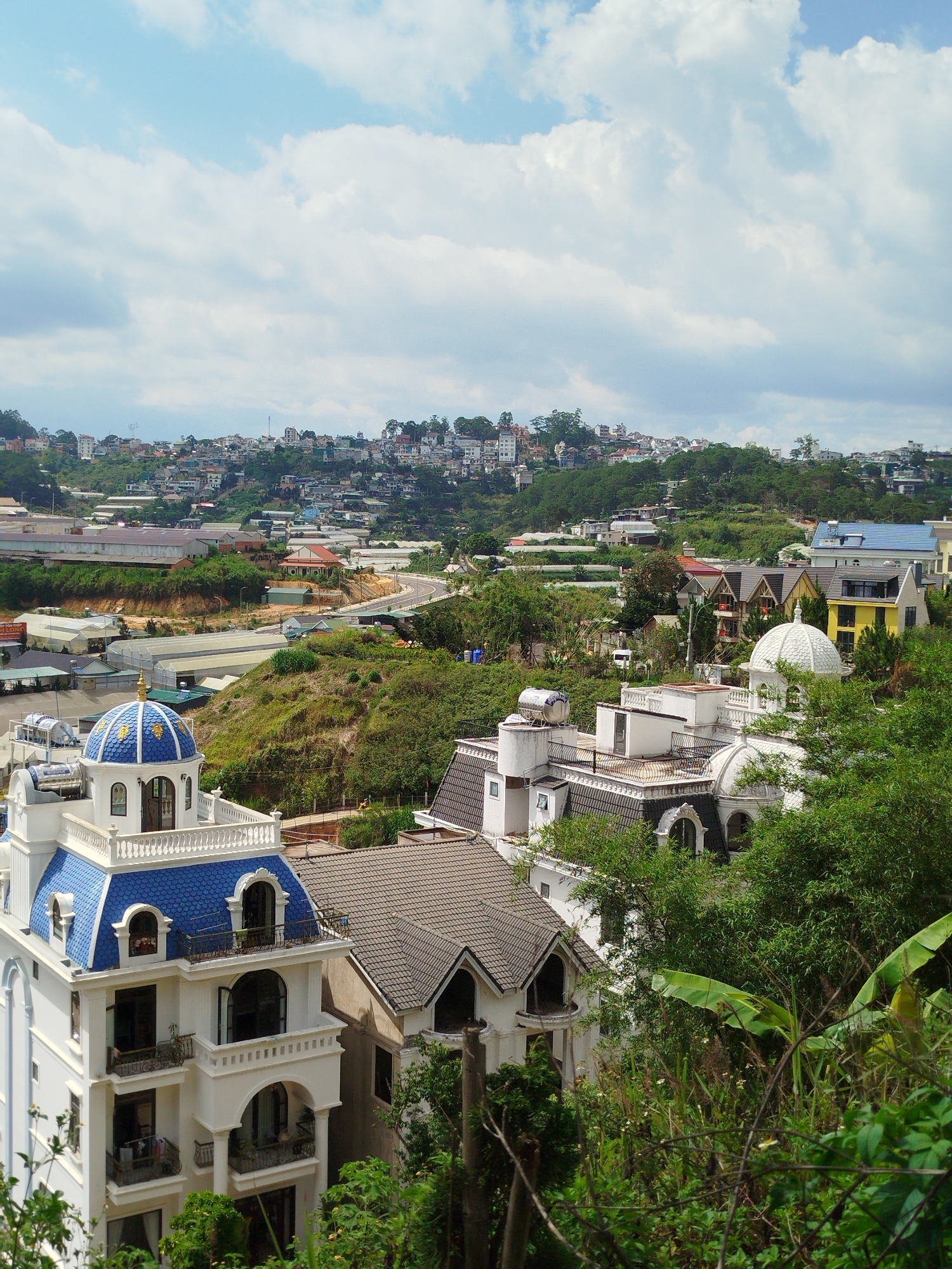 The image depicts a cityscape in Da Lat, Vietnam, characterized by hilly terrain dotted with buildings and lush greenery. In the foreground is a multi-story white building with a distinctive blue-tiled dome and balconies, partially obscured by vegetation. Adjacent to it is a building with a grey roof. The midground showcases more buildings scattered across the landscape, interspersed with trees and foliage. A road winds through the area, connecting different parts of the city. In the background, a densely populated hillside is visible, with numerous buildings clustered together. The sky is partly cloudy with a mix of blue sky and white clouds, adding to the overall brightness of the scene.