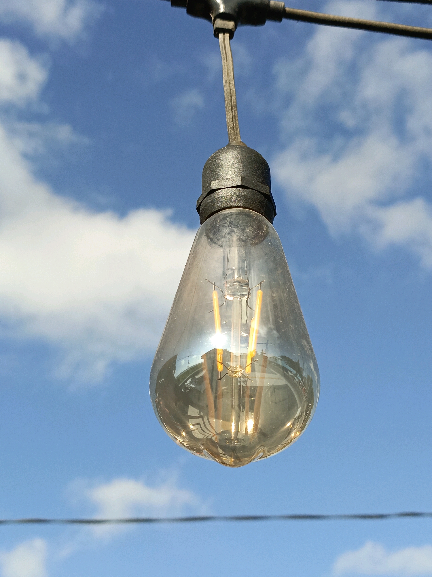 A low-angle close-up shot captures an illuminated lightbulb hanging against a bright blue sky with scattered white clouds. The lightbulb, an Edison-style incandescent bulb with two visible filaments, is suspended from a dark fixture attached to a wire. The glass of the bulb is slightly dusty, and a reflection of what appears to be buildings or structures is visible on its surface. Below the bulb, another dark wire runs horizontally across the frame. The scene is set outdoors, under a clear sky, suggesting a daytime setting. There are no people or animals visible in the image. The image does not contain any specific visual cues that would identify its location within the city of Bulukumba, Indonesia.