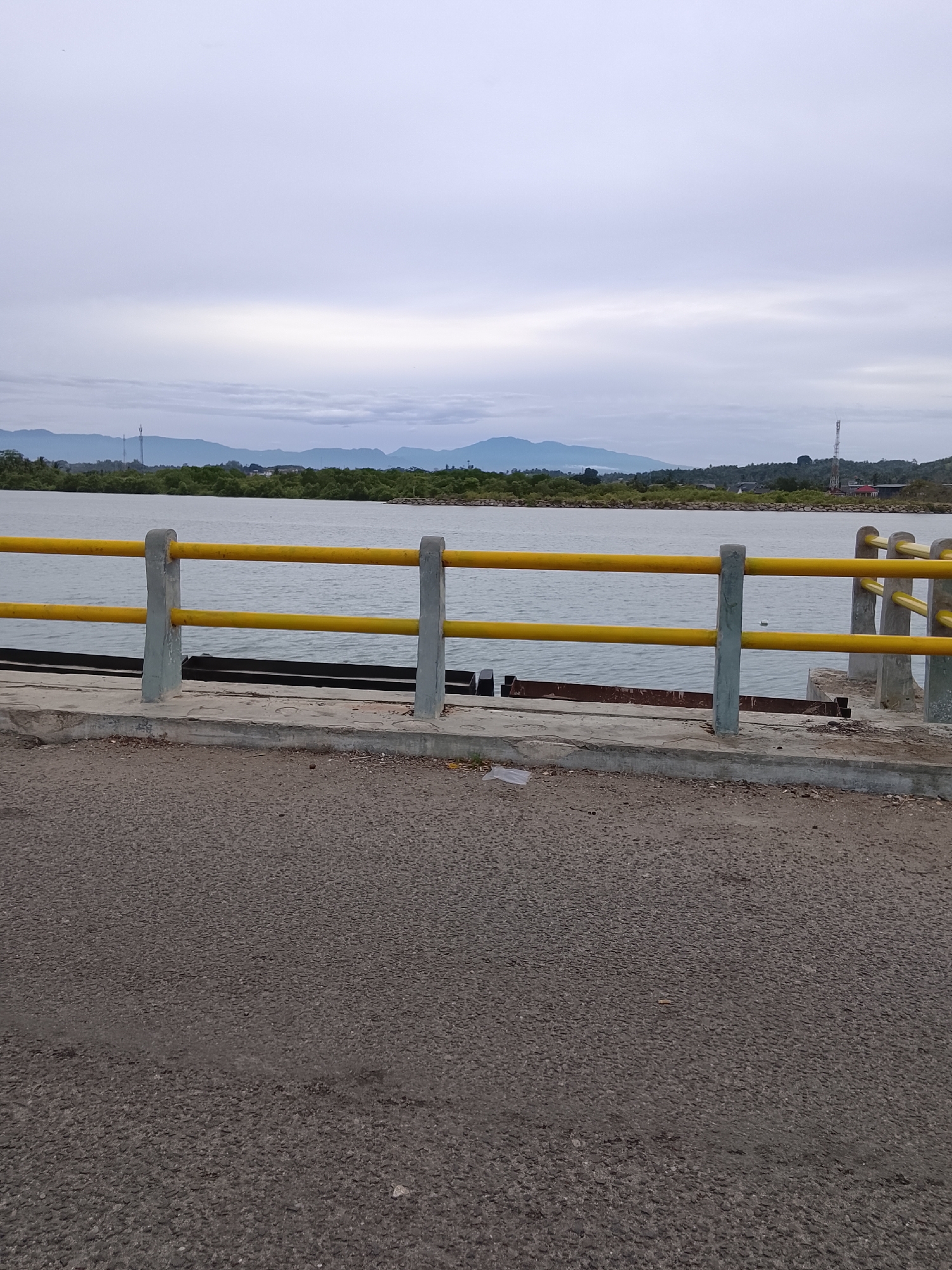 The image shows a view from a bridge in Lhokseumawe, Indonesia. In the foreground is a section of asphalt road, followed by a concrete base supporting a yellow metal railing. Beyond the railing is a body of water with land and mountains visible in the distance under an overcast sky.
