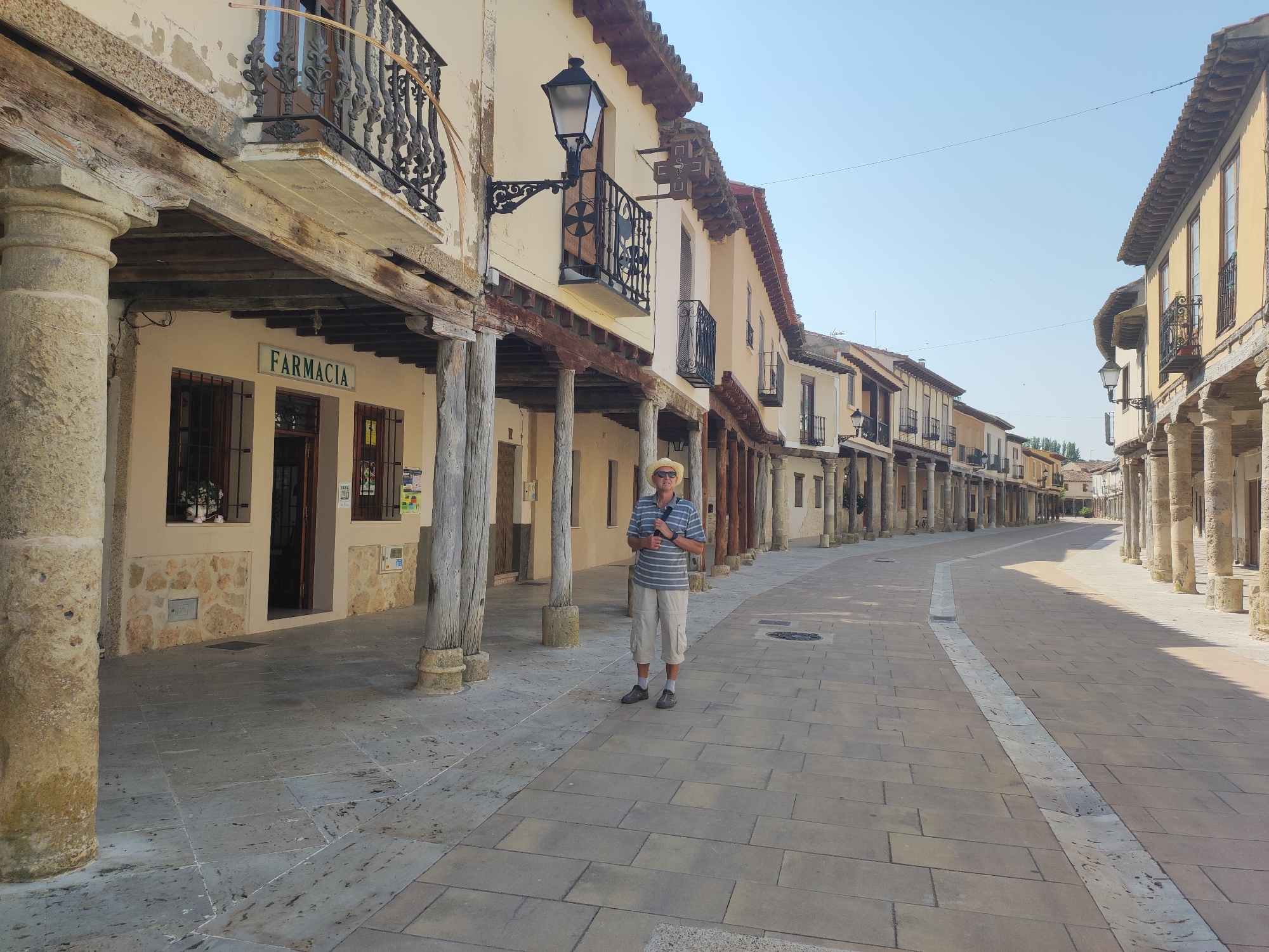 A man stands on a cobblestone street in Ampudia, Spain. He is wearing a straw hat, a striped t-shirt, and beige shorts. He has a bag slung over his shoulder. The street is lined with buildings that have arcades with wooden columns. The buildings have balconies with wrought-iron railings and tiled roofs. The sky is clear and blue. The scene appears to be a historical district.