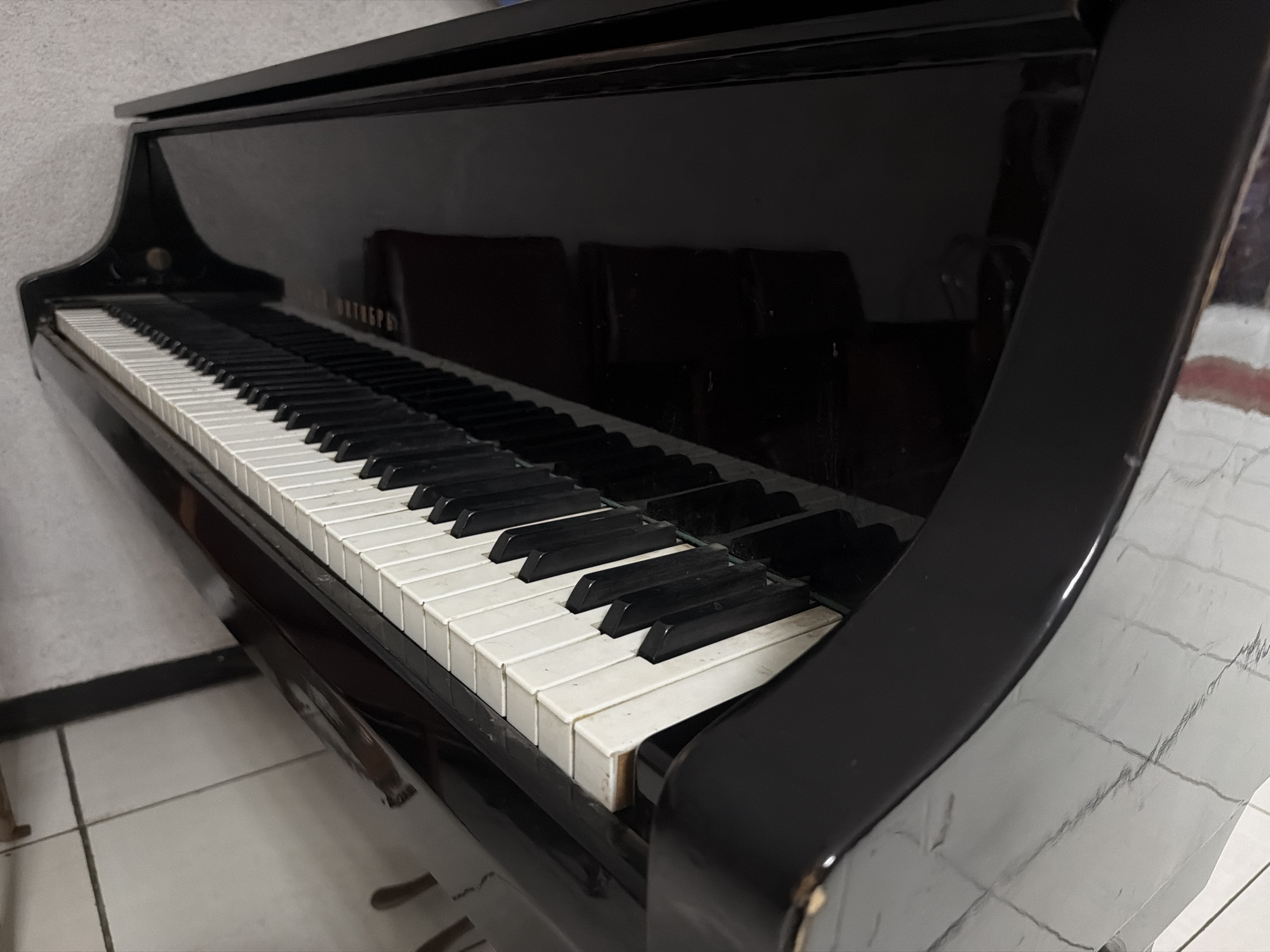 The image displays a close-up, angled view of a black, glossy upright piano in an indoor setting. The piano's highly reflective surface dominates the center and right side of the frame, showing indistinct reflections of the room, including what appear to be dark shapes of furniture or openings.
The piano's keyboard is prominent, featuring white and black keys. The white keys, particularly those in the foreground, show signs of use, with some discoloration and slight chipping on their edges. The black keys also appear somewhat worn. The top lid of the piano is slightly open, revealing another black surface underneath.
To the left of the piano, a light-colored, textured wall is visible. Below the piano, a tiled floor with light-colored square tiles and dark grout lines extends into the foreground and bottom left corner.
Reflected on the fallboard (the cover over the keys), a word written in Cyrillic script is discernible: "ОКТЯБРЬ" (OKTYABR), which translates to "October." This is likely the brand or model name of the piano. No people are present, and no activity is taking place. The overall impression is of an instrument in a domestic or institutional interior.