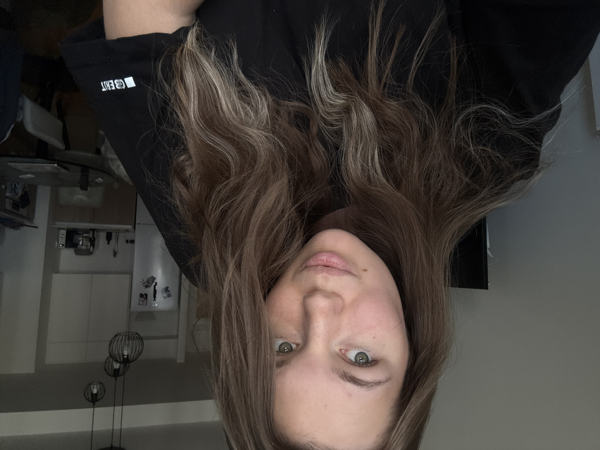 A young woman with long, wavy brown hair and green eyes is taking a selfie in what appears to be an apartment kitchen. She is wearing a black t-shirt with a white logo on the sleeve that reads "GB ERIT". The background shows kitchen cabinets, a refrigerator with magnets, a microwave, a sink, and a dining table with chairs. Three pendant lights hang from the ceiling. The overall lighting suggests it is daytime, but the interior is somewhat dim. The woman is looking directly at the camera with a neutral expression. The location is not specified in the image or its crops, and therefore cannot be determined to be in Belgrade, Serbia.
