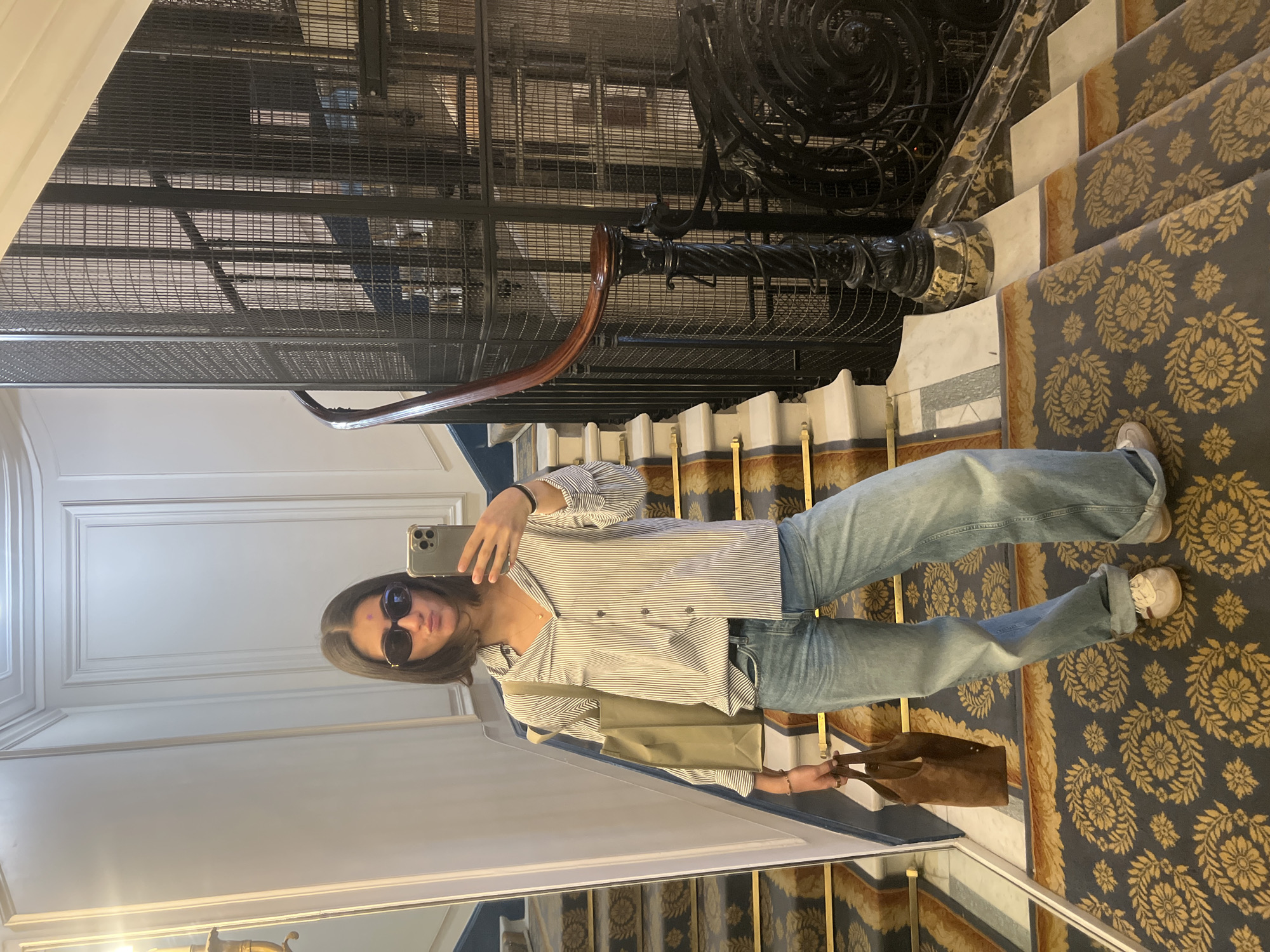 A person is captured taking a mirror selfie on an elegant staircase within what appears to be an old, possibly Parisian, building. The subject, viewed from the chest down, is wearing dark sunglasses, a long-sleeved shirt with thin vertical dark stripes on a white background, light blue wide-leg jeans, and white sneakers. She holds a smartphone, likely an iPhone with multiple rear cameras, in her left hand to take the photo, while an olive green or beige tote bag hangs from her left arm. A brown tote bag is held in her right hand.
The staircase is ornately designed, featuring a dark grey carpet with a repeating gold floral and vine pattern. The risers are white with gold trim, and the banister is made of dark, highly decorative wrought iron with swirling patterns, topped by a polished wooden handrail. In the background, reflected in the mirror, is a towering structure composed of black metal mesh, resembling an elaborate elevator shaft or an internal cage-like staircase. The ceiling visible in the reflection is white with classic decorative molding. The lighting suggests it is daytime. No explicit text is visible.