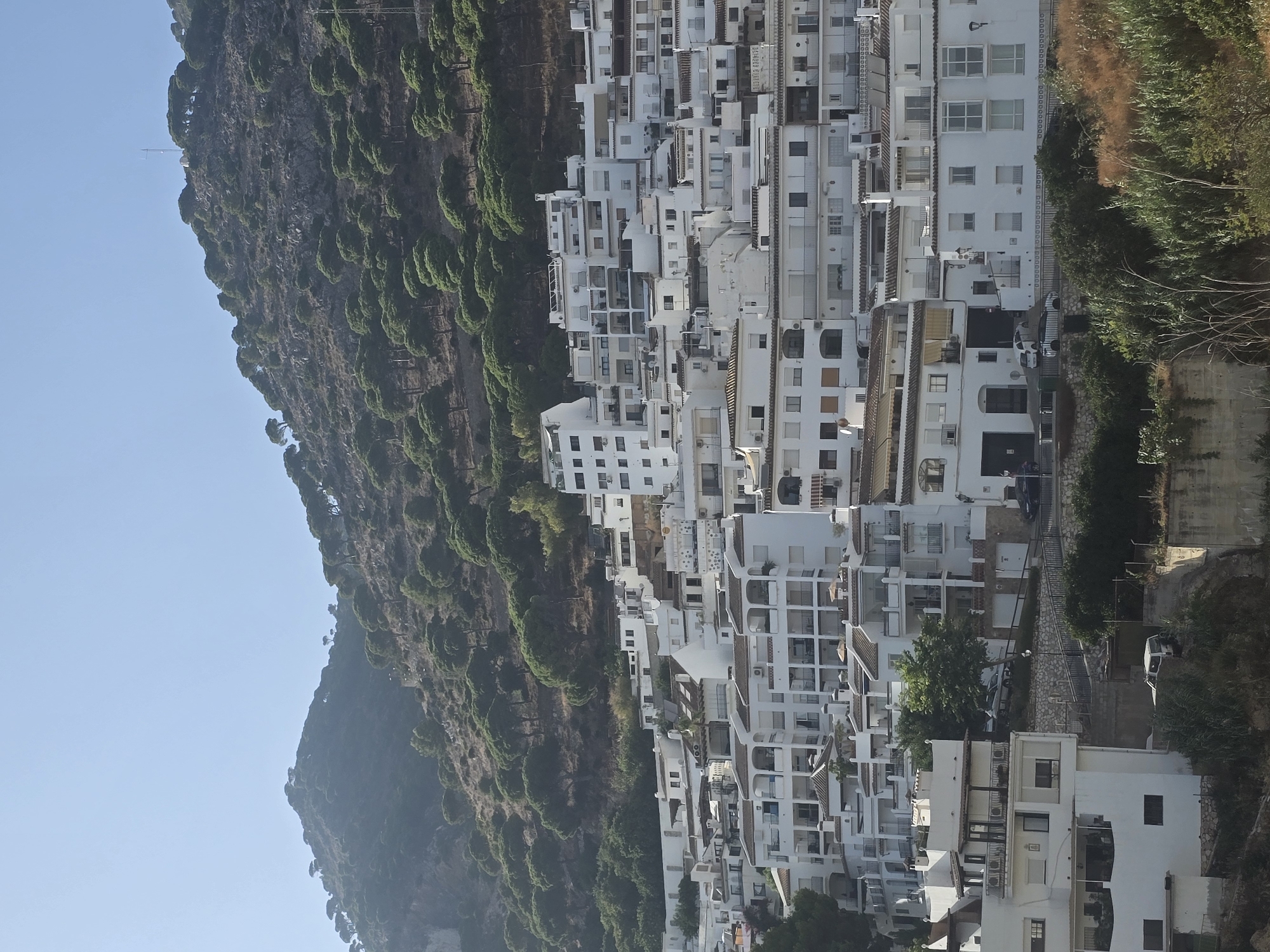 The image depicts the whitewashed houses of Mijas, a pueblo blanco in Andalusia, Spain, cascading down a hillside under a clear blue sky. The buildings are densely packed, with many featuring red-tiled roofs and balconies. Pine trees dot the steep, rocky slopes above the village. A few cars are visible on the narrow streets winding through the town, and some vegetation grows at the base of the hill. The scene is bathed in sunlight, creating a bright and airy atmosphere typical of the Mediterranean.