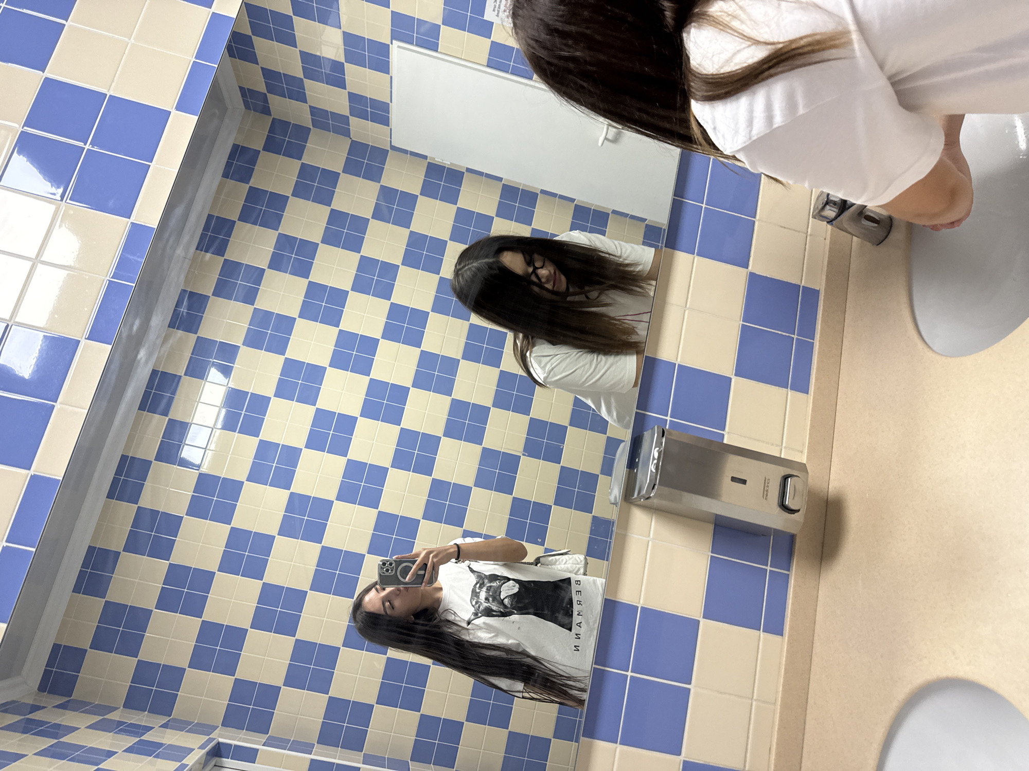 In a public restroom with walls tiled in a blue and cream checkerboard pattern, a young woman with long dark hair takes a selfie in a large mirror. She is wearing a white t-shirt with a black dog graphic and is holding a smartphone to take the picture. Her reflection is visible in the mirror, along with another reflection of her looking down with glasses on. The restroom features a beige countertop with sinks, a silver soap dispenser mounted on the wall, and a white door in the background. The scene is set in Bardar, Moldova.