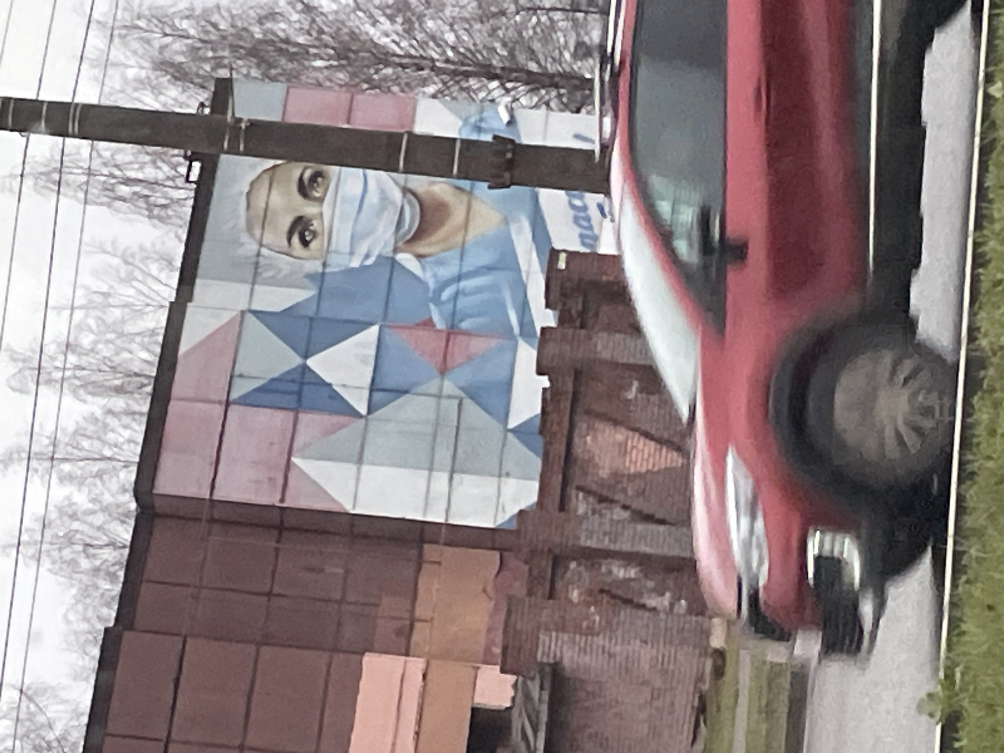 The image shows a mural of a person wearing a surgical mask and scrubs on a building in Izhevsk, Russia. The mural is partially obscured by a red car parked in front of the building. The building appears to be made of brown brick, and there is a telephone pole running vertically in front of the mural. Bare trees are visible in the background. The photo appears to have been taken in the daytime.