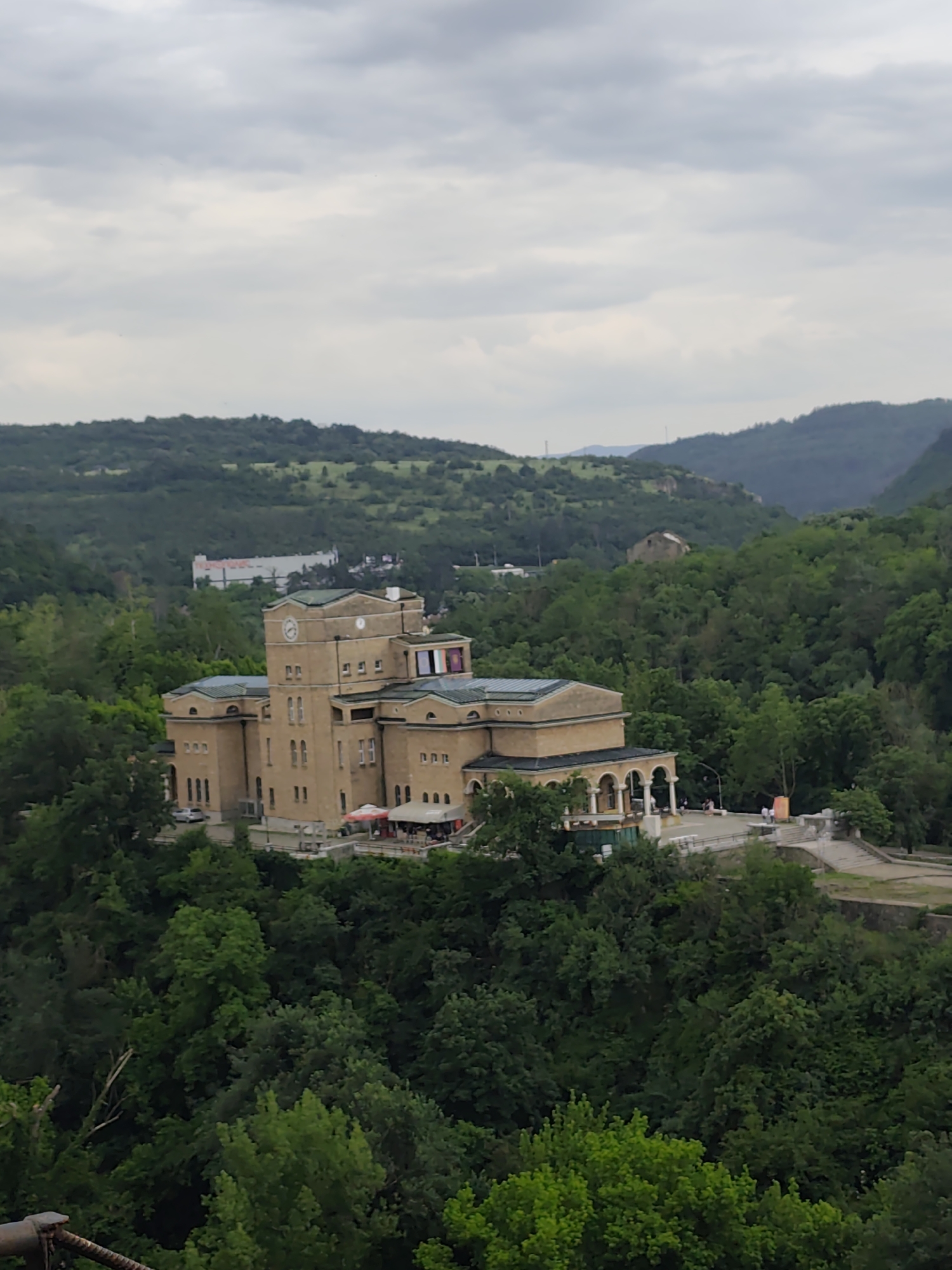 Captured from a high vantage point, the image shows a grand building situated amidst dense greenery under an overcast sky. The building is constructed in a classical architectural style, with symmetrical facades, arches, and a prominent clock tower. A red umbrella can be seen outside of a first story terrace. The surrounding environment consists of thick forests and rolling hills in the background. The location appears to be a city, although the specific location or city is not discernible from the image.
