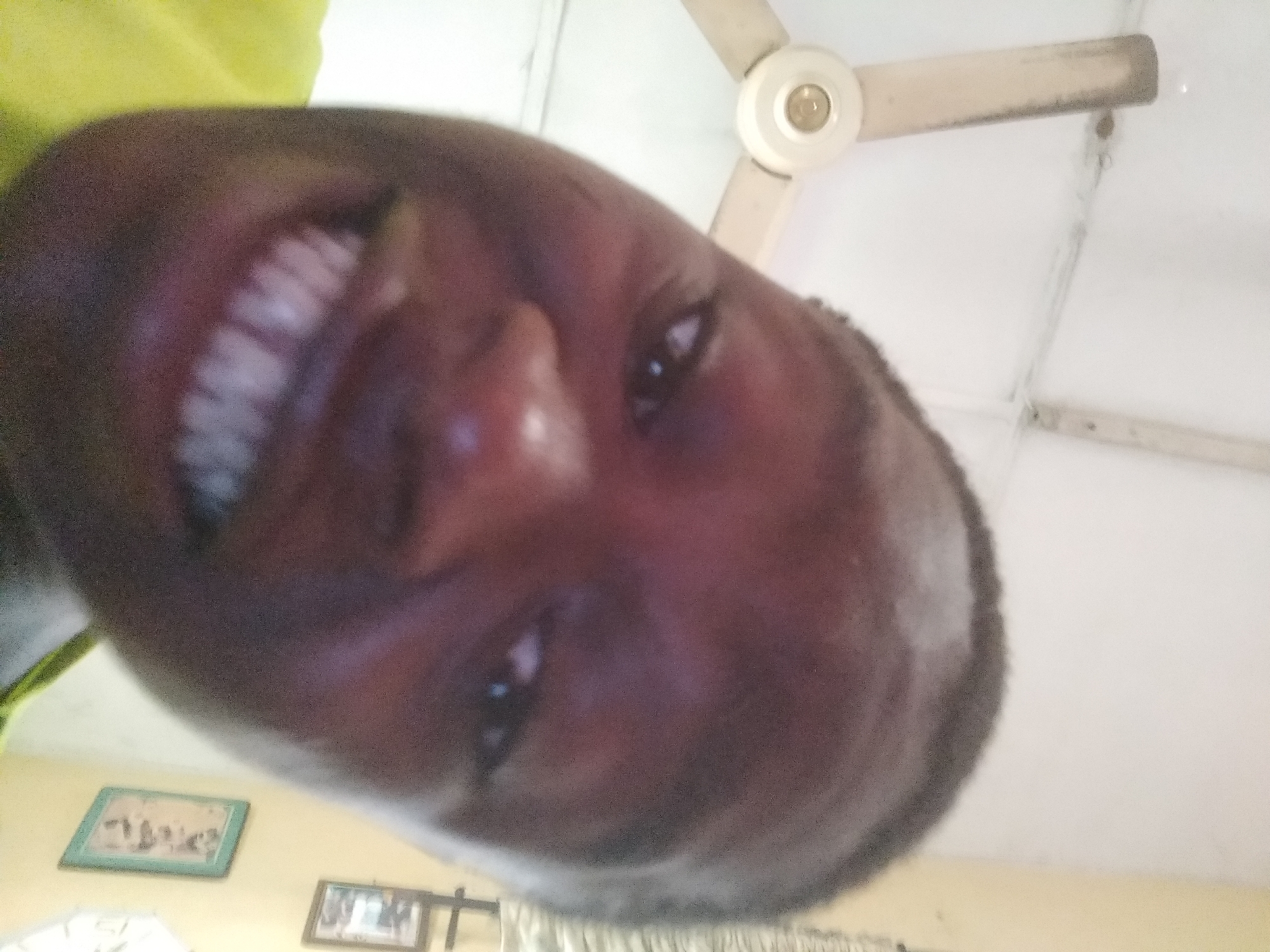 A close-up, tilted portrait of a person smiling, with the camera positioned below and to the side of their face. The person has dark skin and white teeth. In the background, there is a ceiling fan with three blades, and two framed pictures hang on the wall. The location is unknown.