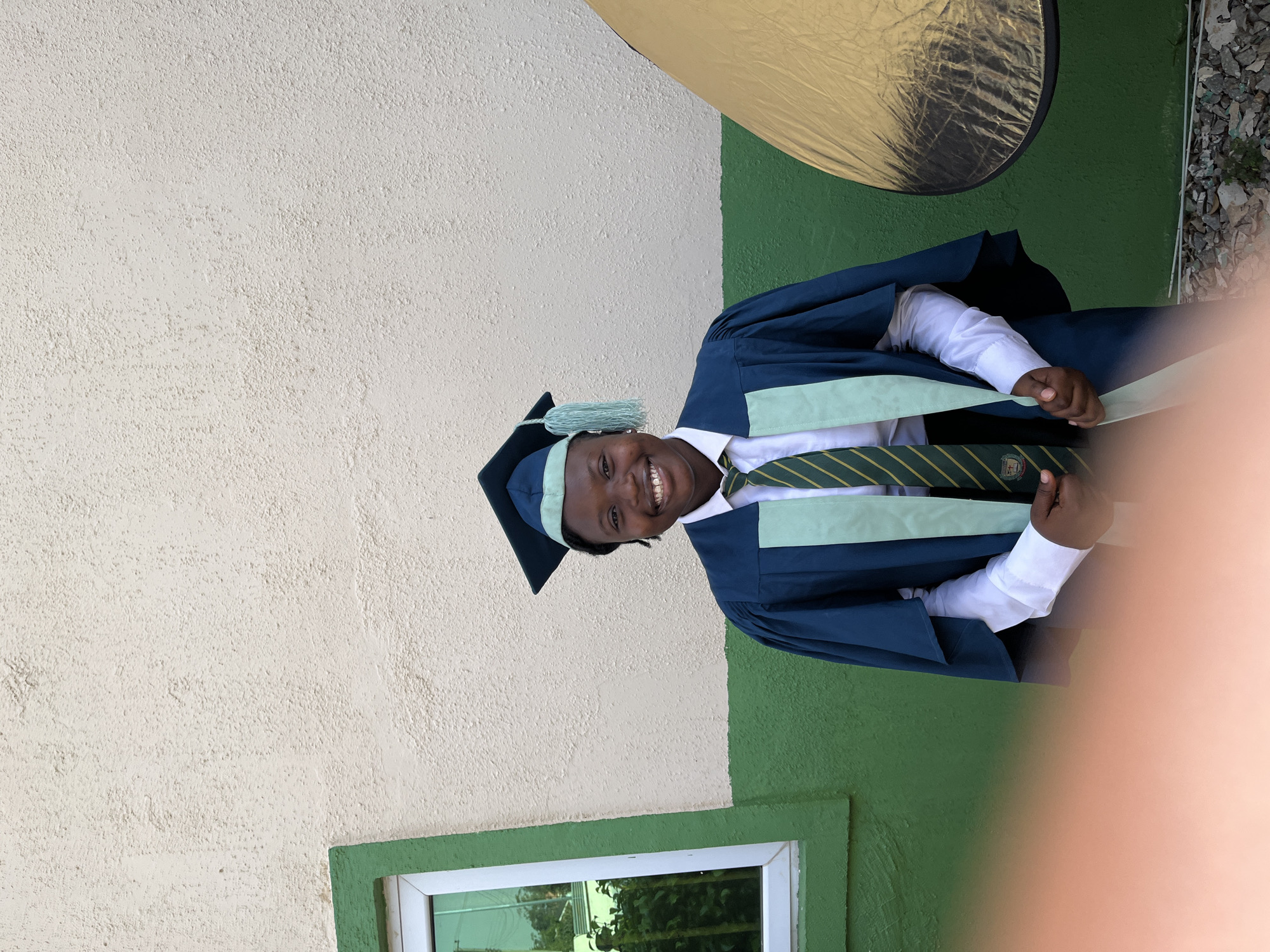 The image shows a person in Jos, Nigeria, wearing a graduation gown and cap. The gown is navy blue with light green accents, and the tie is green and yellow striped. The person is standing against a wall that is painted off-white and green. There is a window with a green frame visible in the bottom left corner. A golden reflector is positioned in the top right of the frame. The ground appears to have some loose gravel and greenery. The person is smiling and holding the light green sash attached to the gown. A partial view of a hand and arm are visible obstructing the right side of the frame.