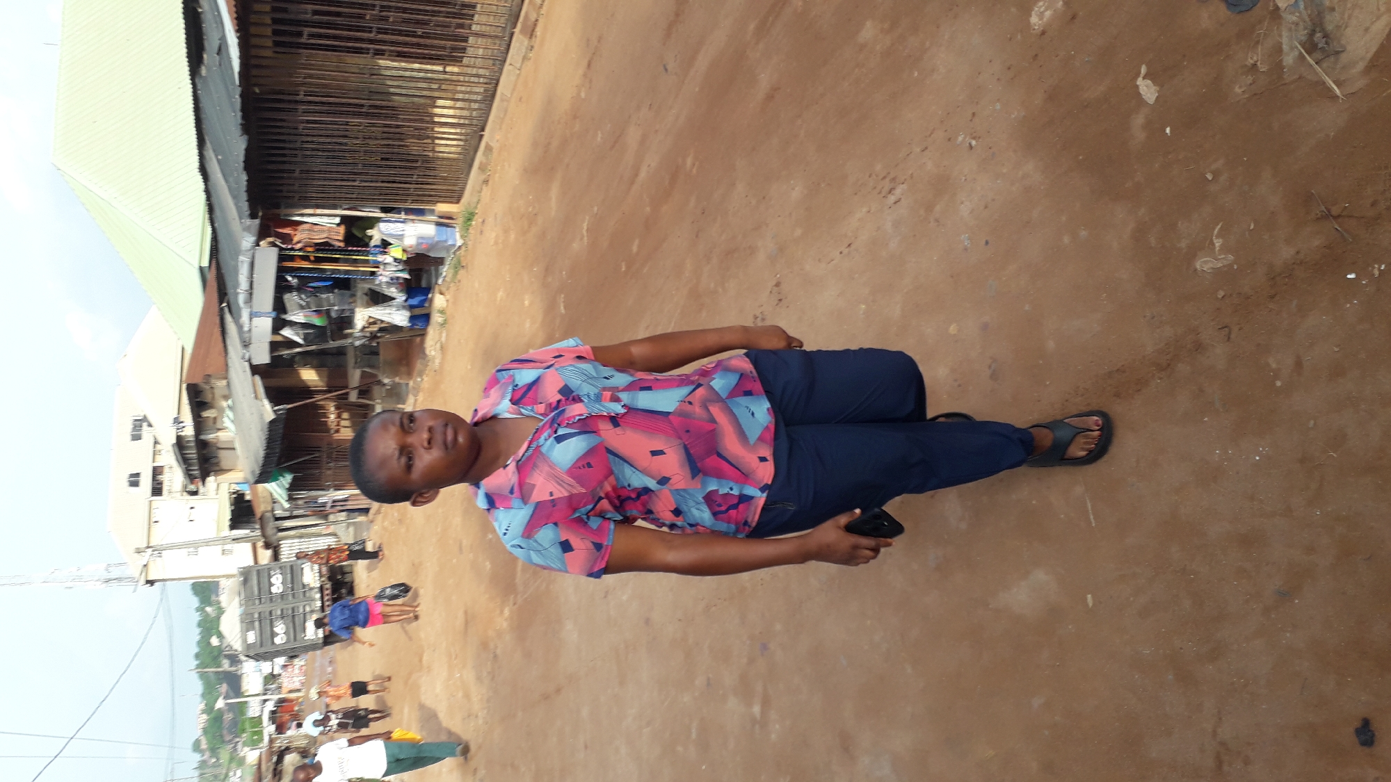 A person stands on a dirt surface in Nkpor, Nigeria. The individual is wearing a short-sleeved shirt with a geometric pattern in shades of pink and blue, dark blue pants, and dark-colored sandals. The person is holding a dark-colored object in their right hand. In the background, a street scene unfolds with buildings, shops, and other people visible. Some of the shops have awnings and closed metal shutters. Other people are walking on the street. The environment appears to be an outdoor marketplace or commercial area.