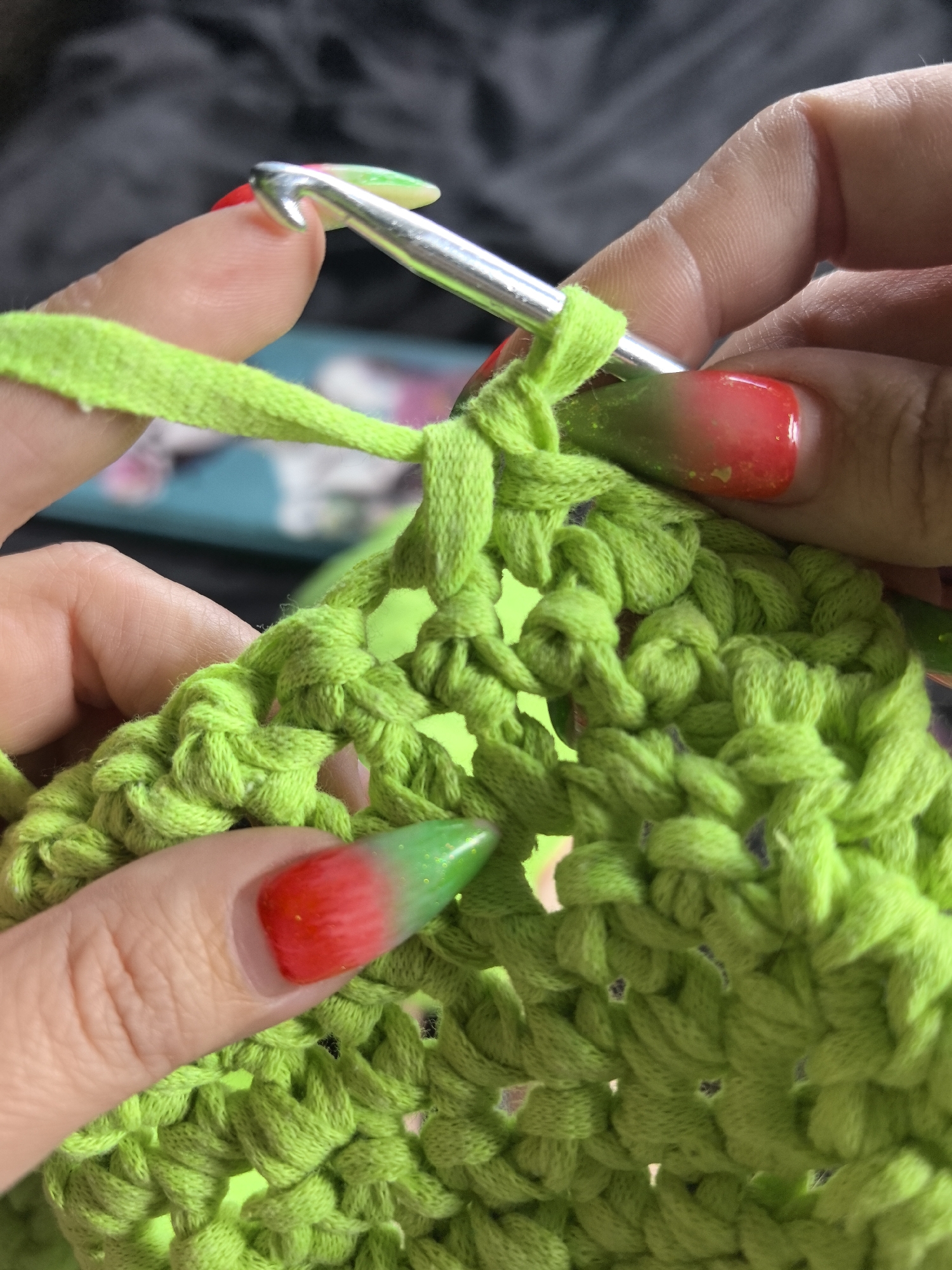 A close-up, high-angle shot captures a person's hands engaged in crocheting with vibrant neon green fabric yarn. The main subject is the activity itself, framed by two hands and the bright green material.
The right hand, seen from the side, holds a silver metal crochet hook. The hook's shaft is inserted into a loop of the green yarn, and a section of its handle, which appears to be a light green and possibly yellow or white plastic, is visible extending upwards. The nails on this hand feature a striking gradient design, transitioning from bright red at the cuticle to a glittery green at the tip.
The left hand supports the evolving crocheted piece, which consists of chunky, loosely-worked stitches made from the same neon green fabric strips. The fingernails on this hand also display the red-to-green gradient with glitter. The yarn itself is not traditional twisted yarn but rather a flat, ribbon-like material, possibly recycled fabric strips or T-shirt yarn, giving the stitches a unique, thick texture.
The background is softly blurred, showing dark, possibly fabric textures directly behind the hands, with a hint of a brighter, teal-colored object further back on the left side. The lighting is bright, highlighting the vivid color of the yarn and the details of the hands and nails. No text is visible in the image. The scene depicts a focused moment of crafting, with the colorful nails adding a personal touch to the activity.