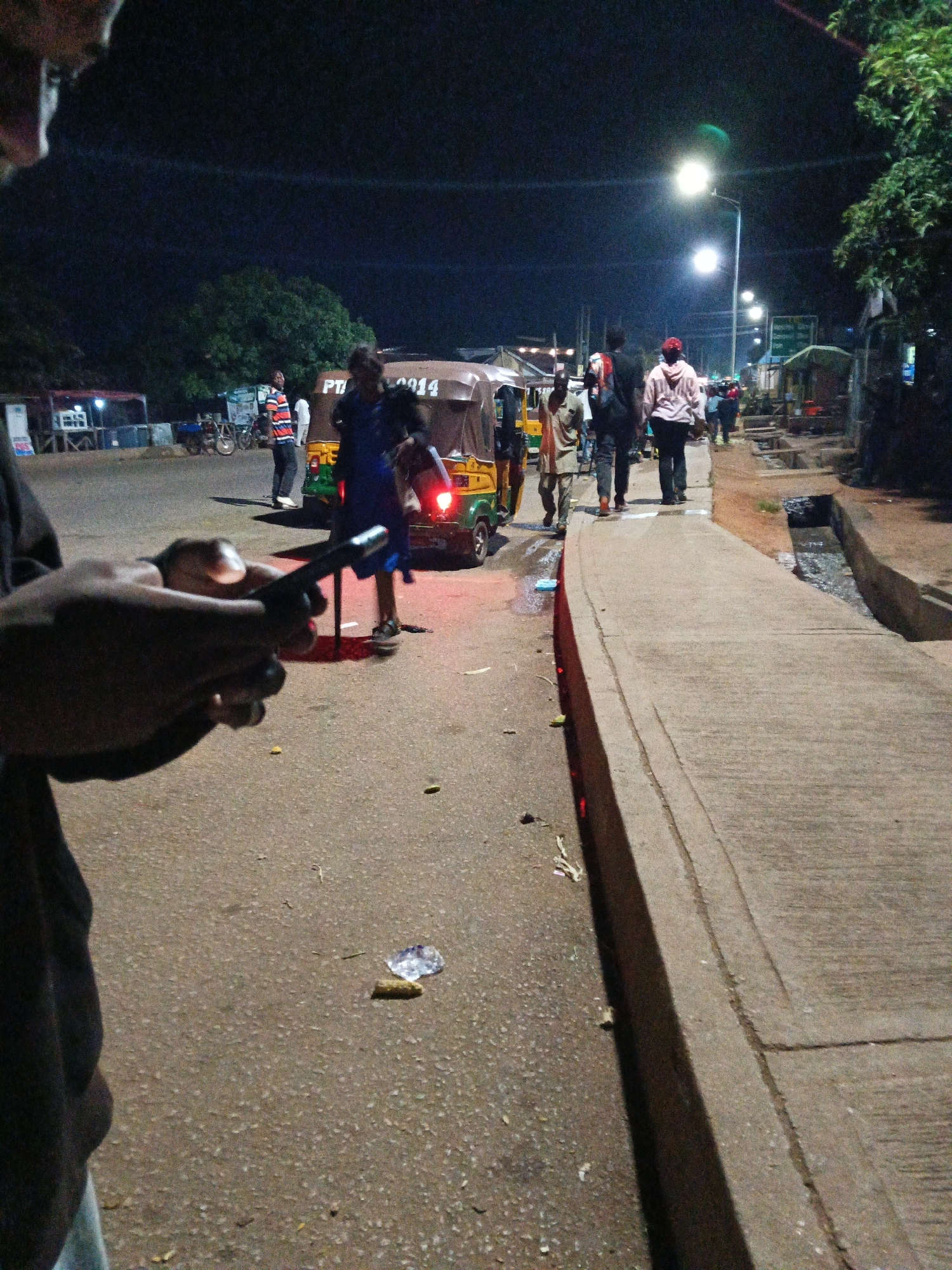 A street scene in Bukuru, Nigeria at night. A person is holding a smartphone in the foreground. In the background, several people are walking on the street and sidewalk. There are also several auto-rickshaws, one of which appears to be a commercial vehicle. Streetlights illuminate the scene, casting long shadows. The street is paved with asphalt, and there is a sidewalk on the right side of the image. The environment appears to be an urban area with some trees and buildings visible.