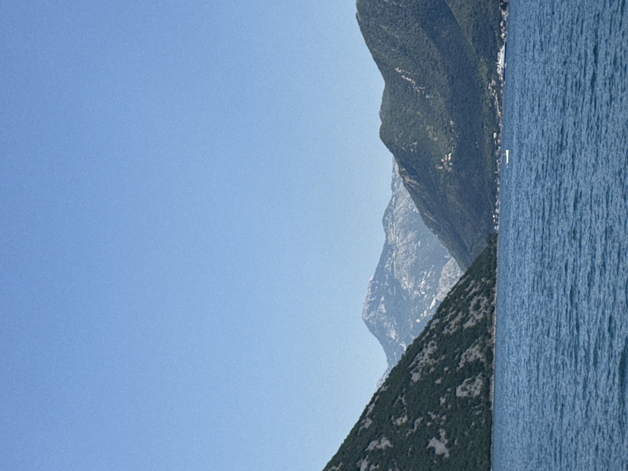 The image depicts a body of water in front of a mountainous landscape. The water is a deep blue with visible ripples. The mountains are covered in green vegetation, and some appear to have rocky, barren slopes. In the distance, a range of lighter-colored mountains with some snow or lighter rock formations is visible. A few small boats are present on the water, and a larger vessel is docked or anchored along the shoreline in the background, near a cluster of buildings that suggest a coastal town. The sky is a clear, light blue. The location is identified as Strp, Montenegro.