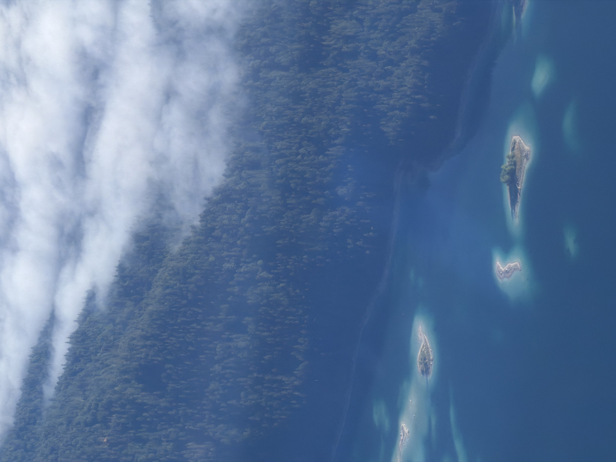 An aerial, wide-angle shot captures a panoramic view of the Eibsee lake in Grainau, Germany. The deep blue water of the lake occupies the lower half of the frame, dotted with several small, tree-covered islands. The larger island in the center is distinctly heart-shaped, with a cluster of trees on its highest point. The upper portion of the image transitions into a dense, dark green forest that carpets a steep mountainside. Wisps of white clouds partially obscure the top of the forest, creating a soft, hazy effect. The overall impression is one of natural beauty, with the serene lake and lush forest contrasting against the ethereal clouds.