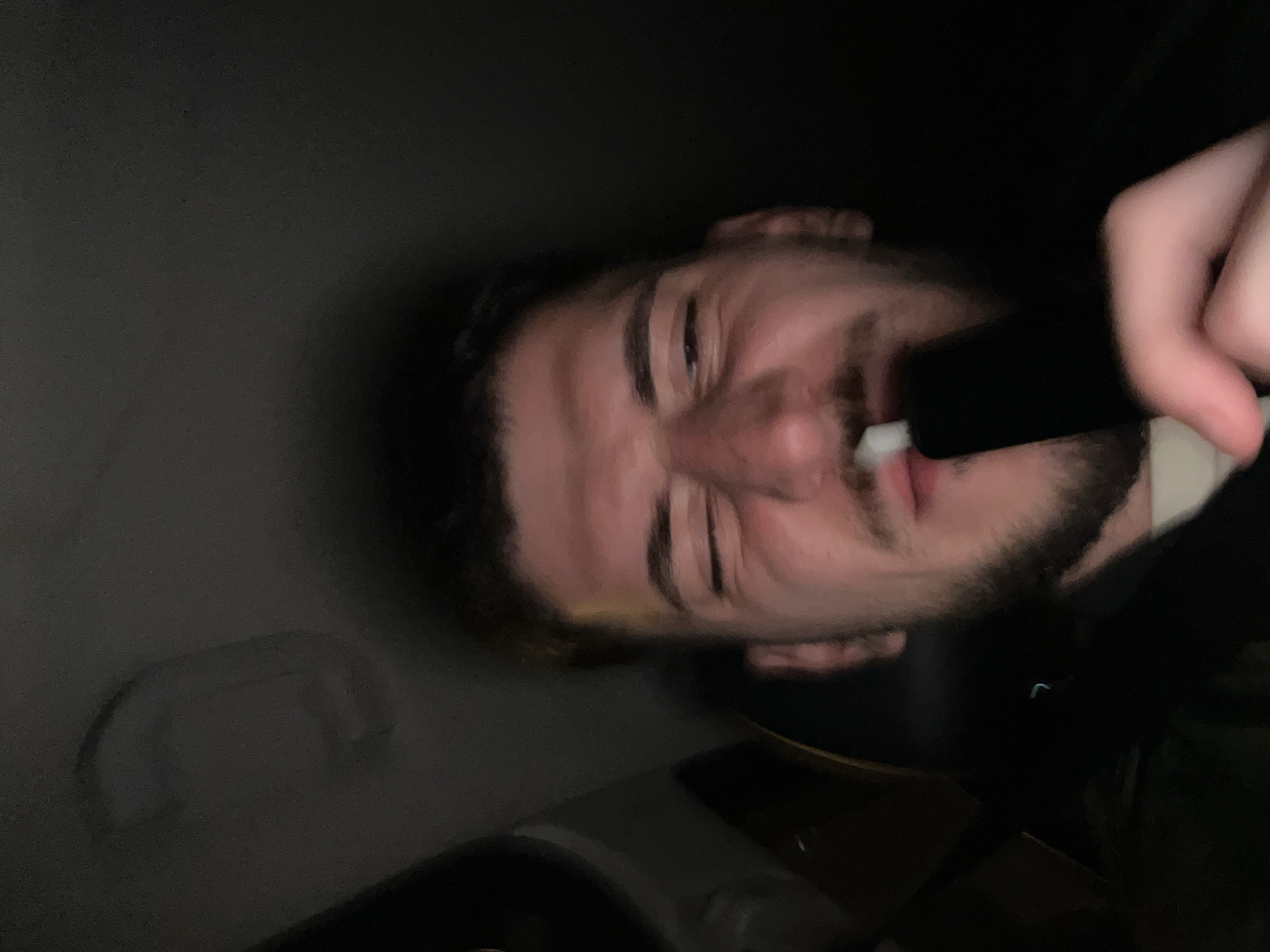 A man with short, dark hair and a beard is holding a black object with a white tip to his mouth. He is squinting his eyes slightly. The background is dark and appears to be the interior of a car, with a handle visible on the left side. The image is rotated. There is no clear indication of Sofia, Bulgaria, or any specific location within the city in the image.