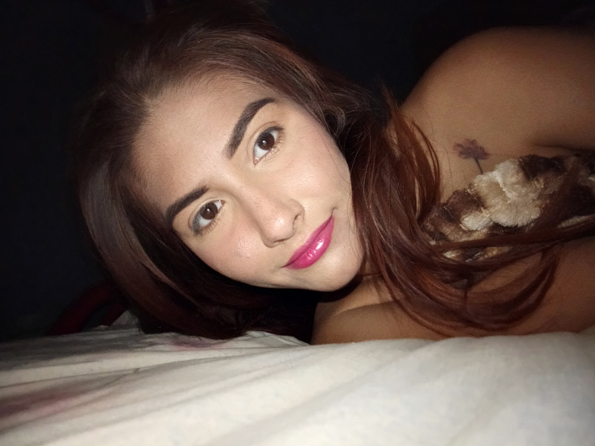 A medium close-up shot shows a woman lying on a white bed. Her head is near the top left corner of the frame, with her body extending diagonally towards the lower right corner. She has brown hair and brown eyes, and she is wearing bright pink lipstick. A tattoo resembling a flower is visible on her upper back, near her shoulder. She is partly covered by a brown and beige patterned blanket. The background behind her is dark, obscuring details. The location is unknown; the description request for Juan Griego, Venezuela cannot be confirmed.