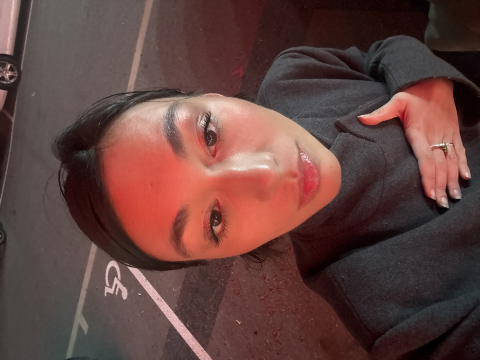 A woman in a gray coat lies on what appears to be asphalt. Her head is angled towards the camera, with a slight upward tilt. She has dark hair and fair skin, accentuated by a red light source. Her hand, adorned with rings, rests on the coat. The background shows a disabled parking symbol, hinting at the setting. The overall composition conveys a sense of casual repose and perspective.