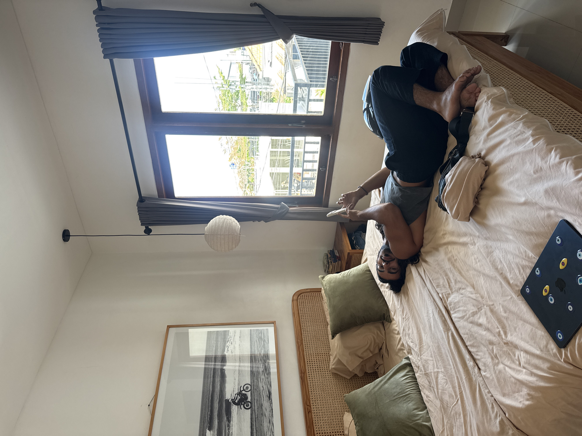A man with dark hair and a beard is shown lying on his back on a large bed, looking towards the camera with a relaxed expression and a slight smile. He is wearing dark shorts and a dark hooded top, with his feet crossed.
The bedroom is bright, with light-colored walls. The bed is covered with a light-colored duvet or sheet and features a light-colored woven headboard. Several pillows are on the bed, including two large light-colored ones and two olive-green velvet-like throw pillows. To the man's right, a dark laptop with multiple round, eye-like stickers (some blue, some yellow) is resting on the bed; an Apple logo is visible beneath some stickers. A light-colored bag with a black strap is also positioned near his feet.
On the left side of the room, a large wooden-framed window allows ample daylight inside. Dark grey Roman blinds are partly drawn. Through the window, glimpses of other buildings and greenery can be seen. Below the window, a large framed black and white print hangs on the wall, depicting a person on a motorcycle in a sandy environment. A spherical, woven pendant light fixture hangs from the ceiling, along with a track light. A wooden shelf with some books is partially visible behind the bed's headboard.
The scene captures a moment of relaxation during the daytime, in a comfortable bedroom setting. The casual pose and relaxed expression of the man suggest contentment. The location is Kuta Selatan, Indonesia.