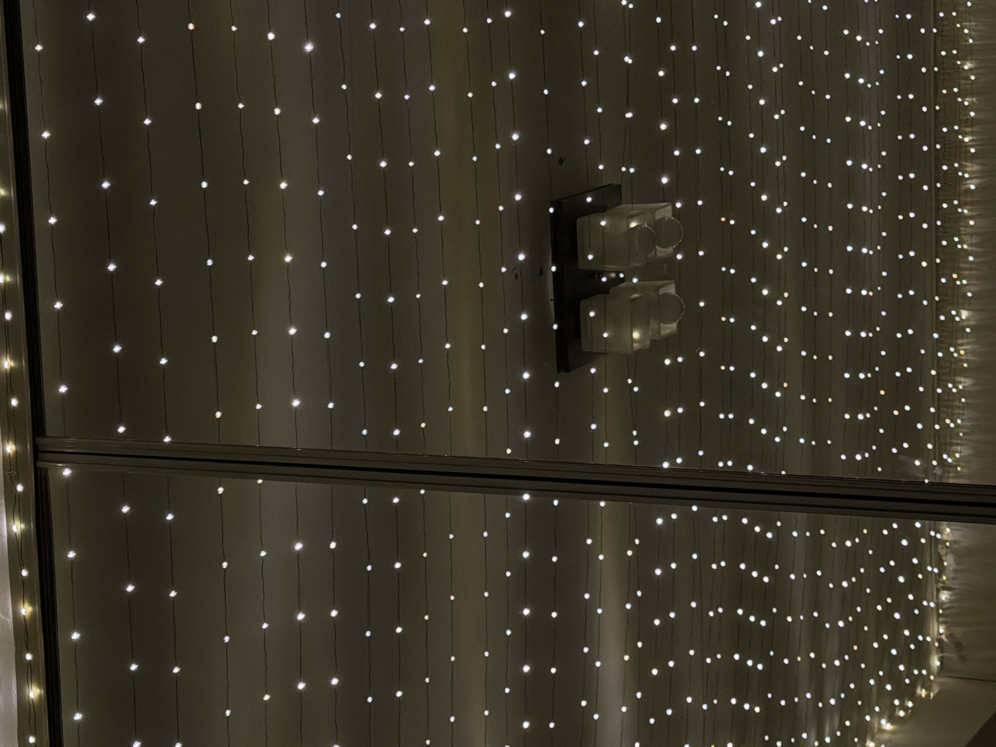 The image shows a close-up view of a wall adorned with strings of lights, resembling a curtain of small, bright points. Positioned on the wall is a light switch with two toggle switches. A reflective border traverses the image, creating a visual division. The location is not discernible from the provided image.