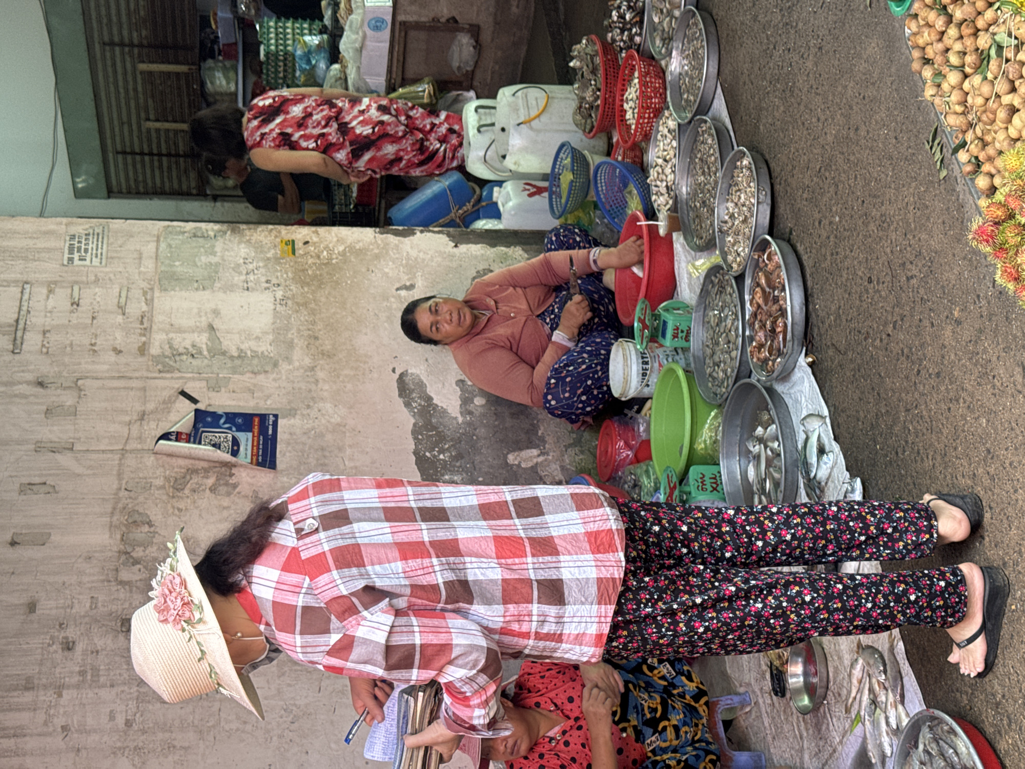 The image depicts a bustling outdoor market scene, likely in Mũi Né, Vietnam, with a focus on fresh seafood for sale. In the foreground, a woman wearing a wide-brimmed straw hat adorned with pink flowers and a red and white checkered shirt stands, holding a stack of documents and a pen. She is partially obscuring another woman in a red polka-dot shirt and patterned pants who is seated on the ground. To the right, a woman wearing a light brown long-sleeved shirt and blue and white patterned pants is seated, actively engaged in preparing seafood, holding a knife. In front of her are various baskets and containers filled with shellfish. The ground is covered with a white tarp, displaying an array of seafood, including fish and shellfish arranged in metal basins and red baskets. In the background, other market stalls and vendors are visible, with a person in a red and white patterned dress standing near shelves stocked with goods. The environment suggests a typical Vietnamese market with a rustic, open-air setup against a weathered wall.