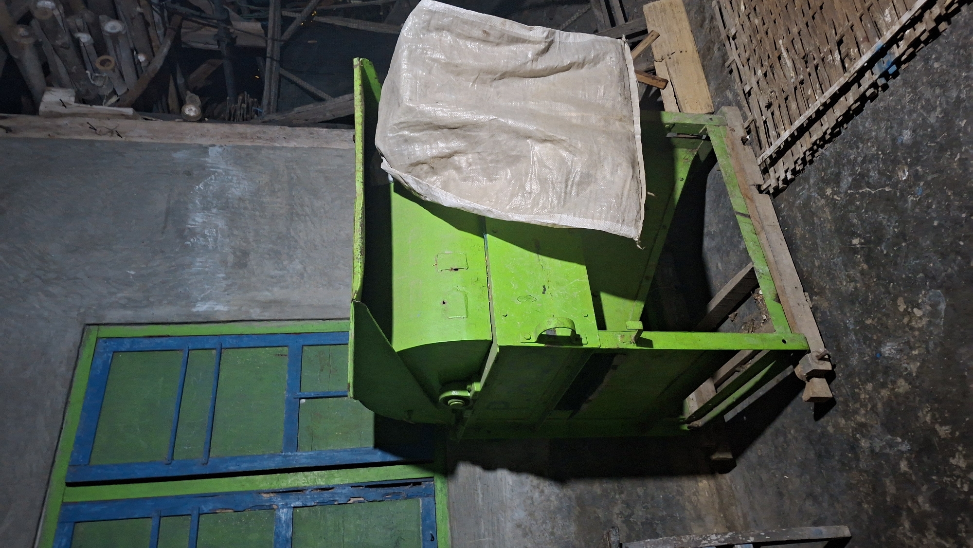 In Sambeng, Indonesia, a bright green mechanical thresher sits on a dark, speckled concrete floor. The thresher, constructed of painted metal, has a large chute at the top and a bag made of white woven material attached to its right side. The machine is supported by a wooden frame. To the left of the thresher, a window with blue and green painted wooden frames is visible against a textured gray wall. To the right of the thresher, wooden planks and what appears to be a woven mat are present on the floor. The overall environment suggests an outdoor or semi-enclosed space, possibly a workshop or agricultural area.