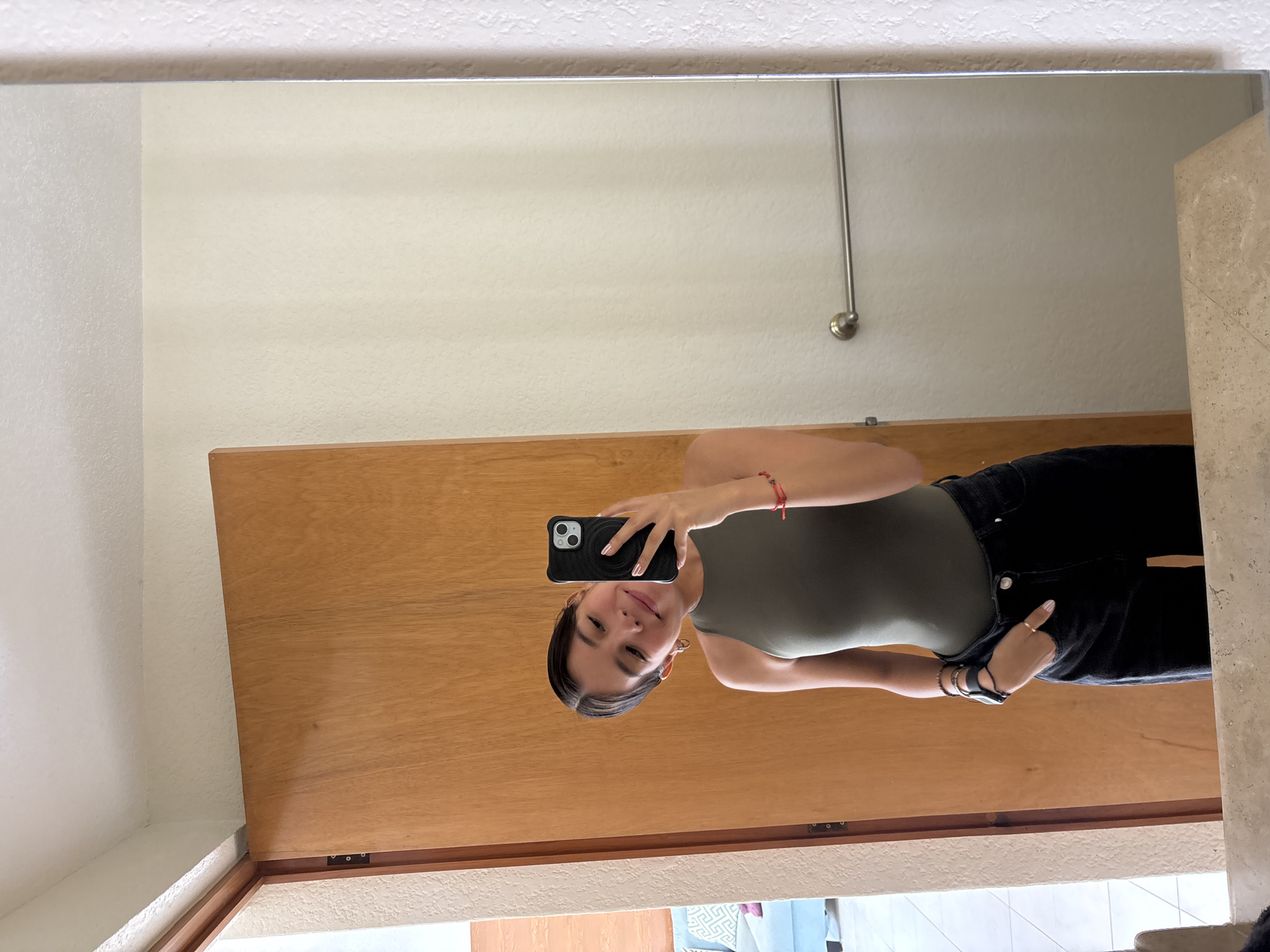 The image displays a mirror selfie, rotated 90 degrees counter-clockwise. The main subject is a young woman, positioned on the left side of the frame as she appears in the mirror reflection. She is holding a dark smartphone with a ring grip in her right hand, using it to take the photograph. She has dark hair, a subtle smile, and is looking towards the phone's camera.
The woman is dressed in a form-fitting dark olive green sleeveless top or bodysuit paired with black pants. She wears a red string bracelet on her right wrist, and multiple bracelets, including what appears to be a smartwatch, on her left wrist, along with a ring on her right hand. Her nails are painted a light shade.
The setting, reflected in the mirror, includes a prominent light brown wooden door positioned behind the woman. To the left and top of the reflected scene, a light-colored, textured wall is visible. A metallic rod or pipe extends vertically downwards from the upper portion of this wall. On the far right edge of the mirror's reflection, a vertical section of a light beige, stone-like structure is present. The lower part of the reflection reveals a light-colored tiled floor with indistinct shapes. The lighting suggests a well-lit indoor environment. No visible text is present in the image. The context provided indicates the location is Puebla, Mexico.